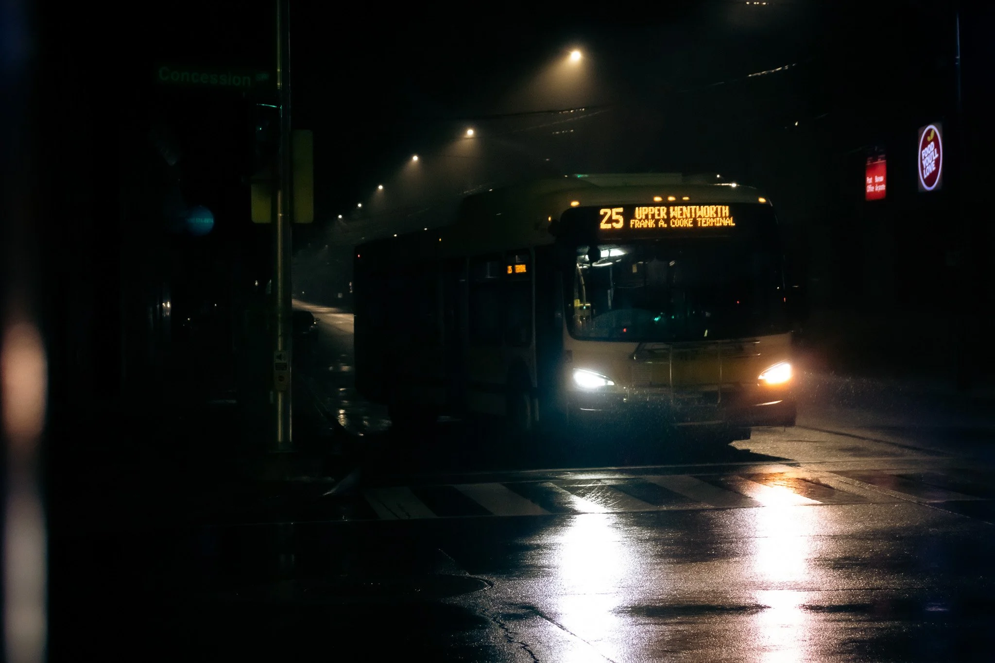 Nighttime scene of a city bus with a sign displaying route 25 to Upper Wentworth, Frank A. Cooke Terminal, on a wet street with reflections, illuminated by streetlights.
