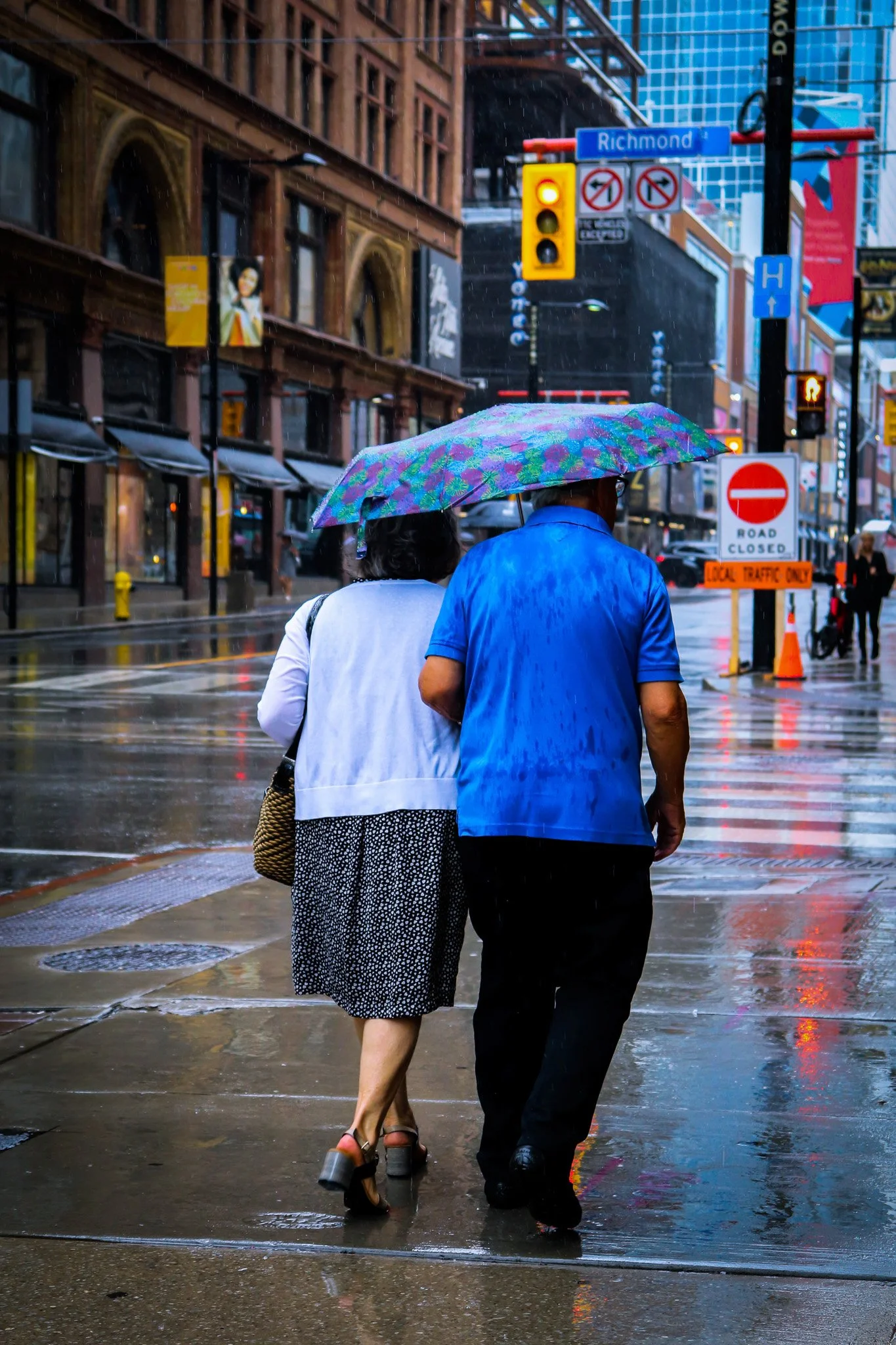 Two people walking together in the rain under an umbrella on a city street, with wet sidewalks and illuminated traffic signals.
