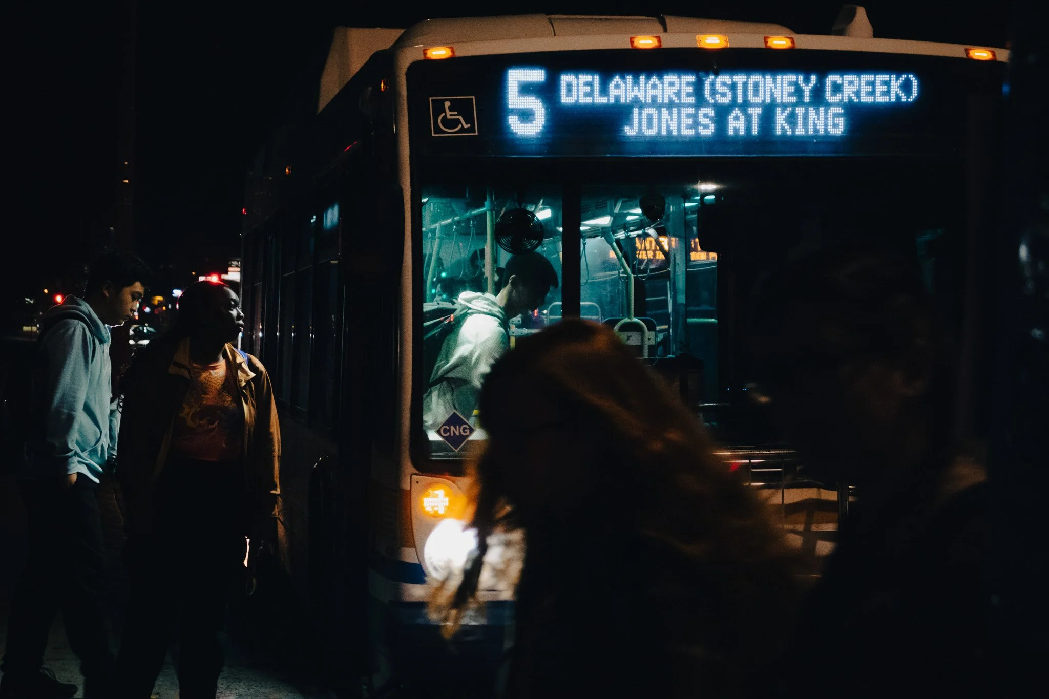 A city bus at night with people boarding and disembarking. The bus displays route number 5 heading to Delaware (Stoney Creek, Jones at King) and has a wheelchair accessibility sign. The scene is dark with minimal lighting.