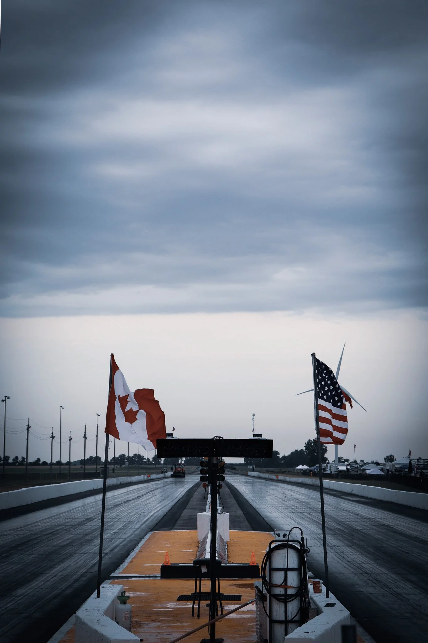 Race track with a starting line and flags of Canada and the United States. Overcast sky, some equipment and cars in the background, with wet asphalt.