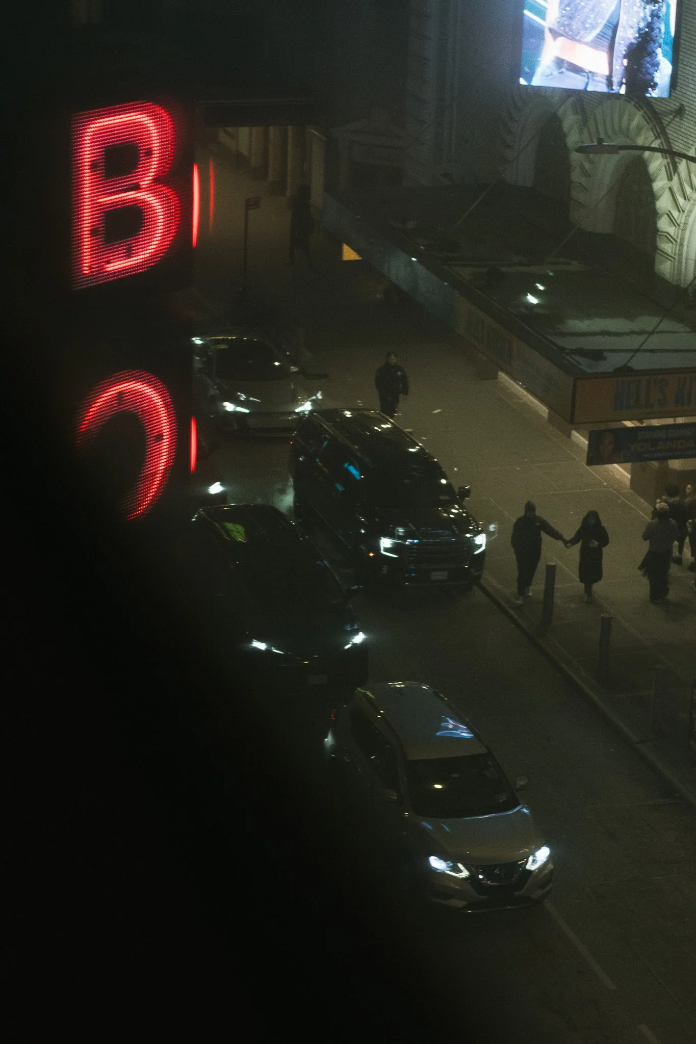 Nighttime scene showing a parking lot outside a building with a large red-lit letter B sign, several cars parked, some with headlights on, and a small group of people holding hands near the sidewalk, with an electronic billboard displaying an adverti