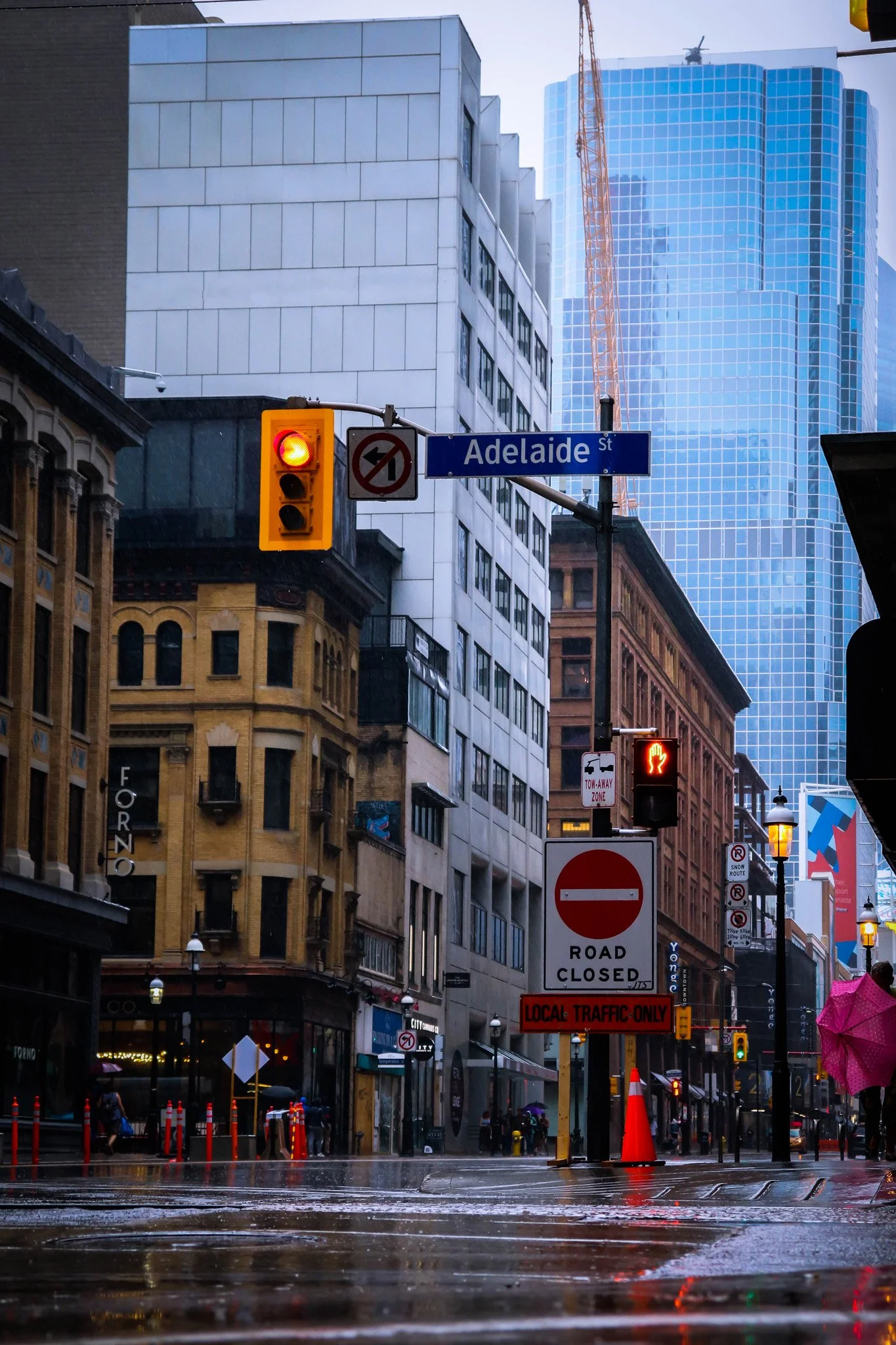 A city street scene during a rainstorm with wet roads, traffic lights, and tall modern buildings in the background, including a skyscraper with glass windows and a crane.