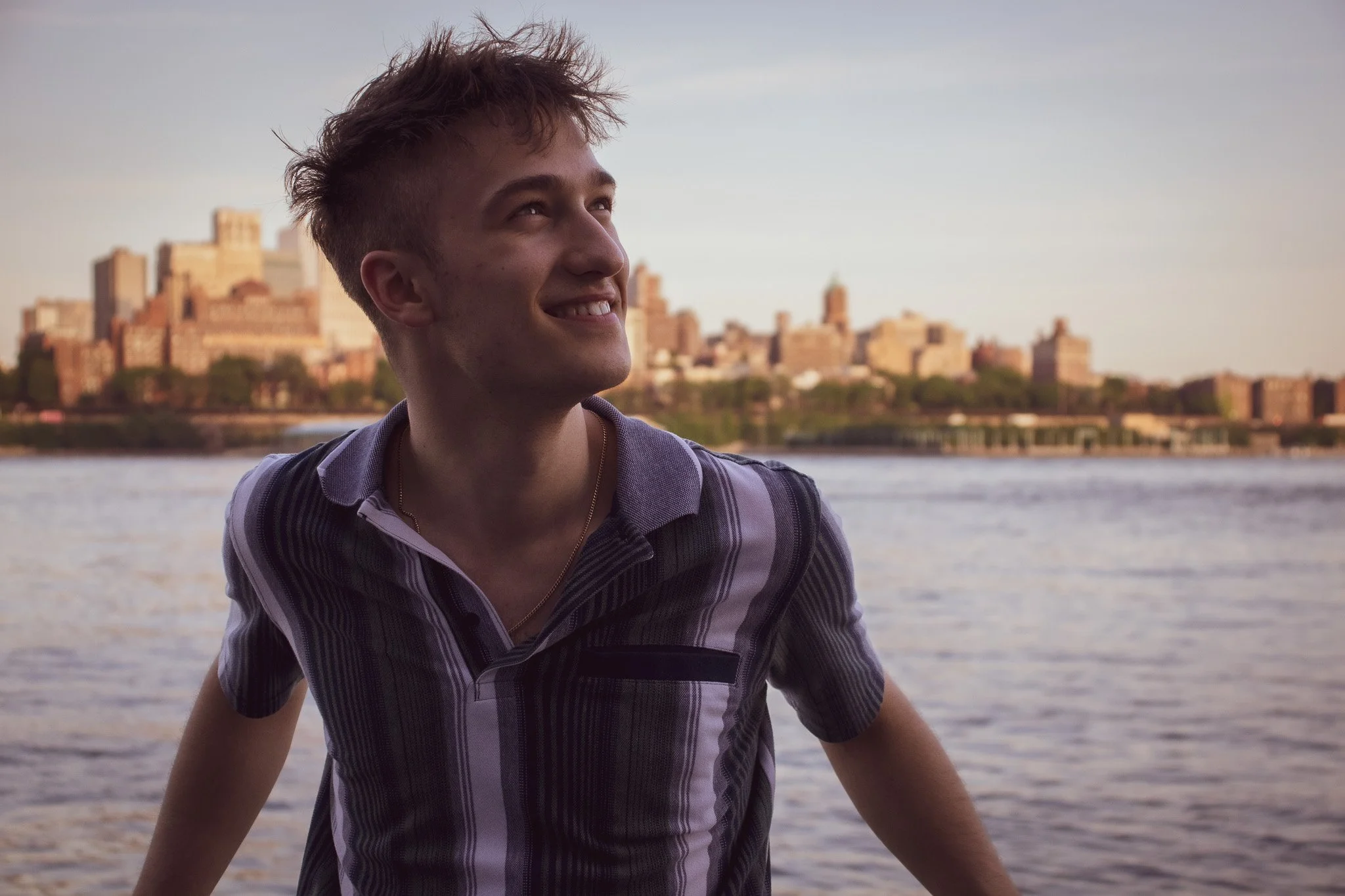 A young man with short, spiky hair smiling and looking up while standing near a body of water with a city skyline in the background during sunset.