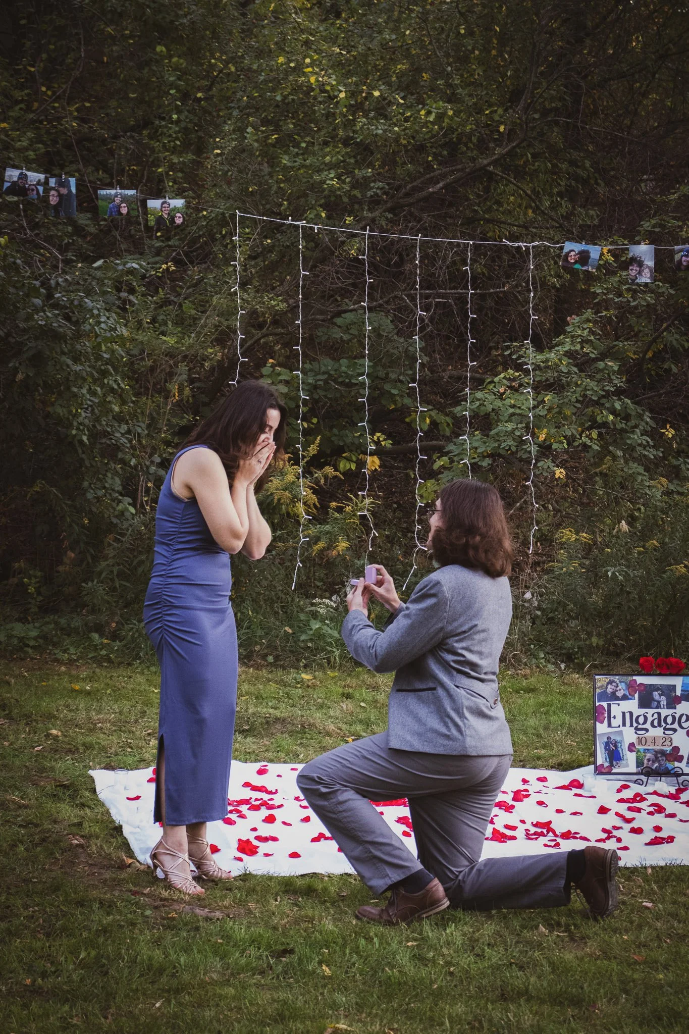 A woman in a blue dress is kneeling and proposing to a woman in a grey dress outdoors on a white blanket decorated with red rose petals. There are photos and a sign that says "Engagement" in the background, along with string lights and a wooded area.