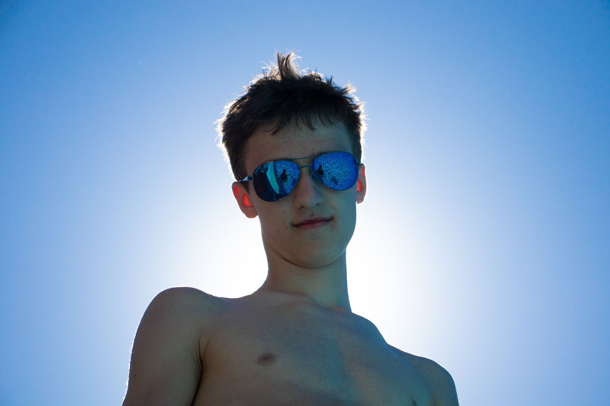 A young person with short, dark hair wearing reflective sunglasses, looking at the camera against a clear blue sky with sunlight creating a backlit effect.