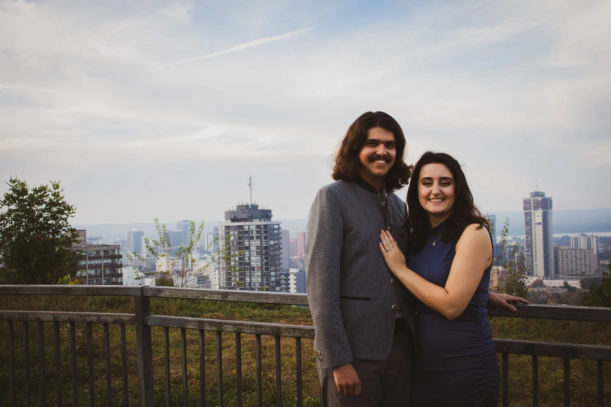 A smiling couple standing outdoors with a city skyline in the background, including several tall buildings and a cloudy sky.
