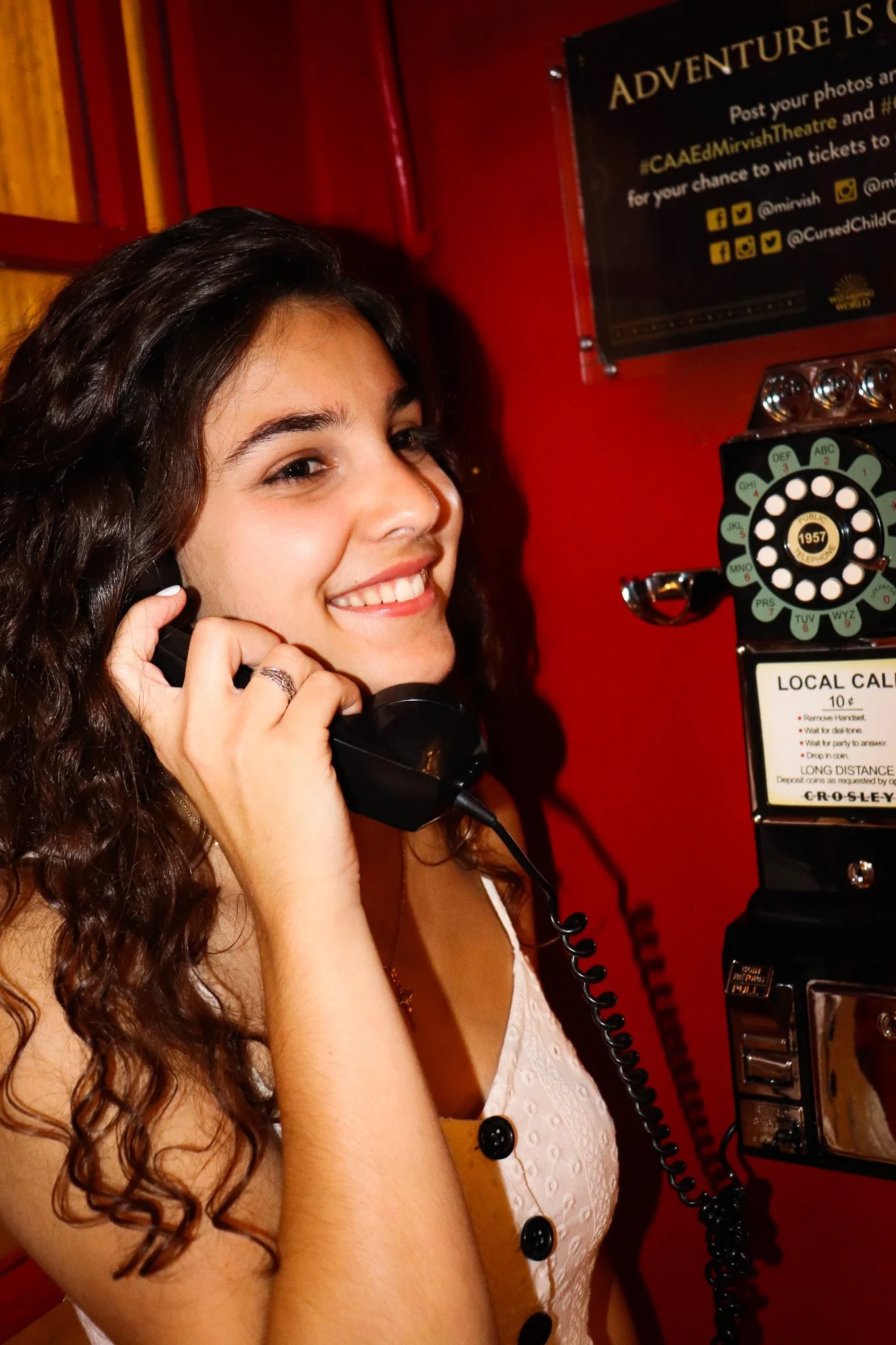 A young woman with long, curly brown hair smiling while talking on a payphone. The background includes a red wall and a black sign with yellow text about posting photos for a contest.
