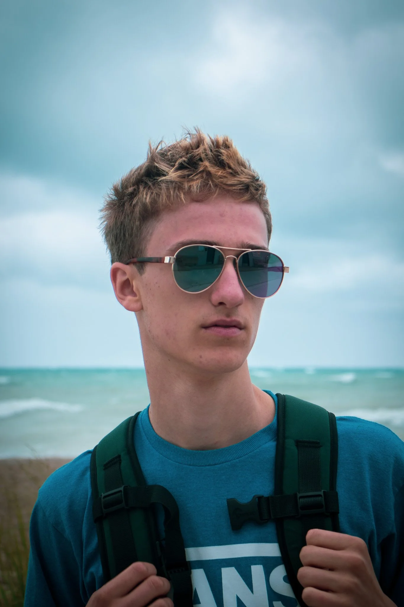 A young man with brown hair wearing sunglasses and a blue T-shirt, holding the straps of his black backpack with both hands, standing on a beach with the ocean and cloudy sky in the background.