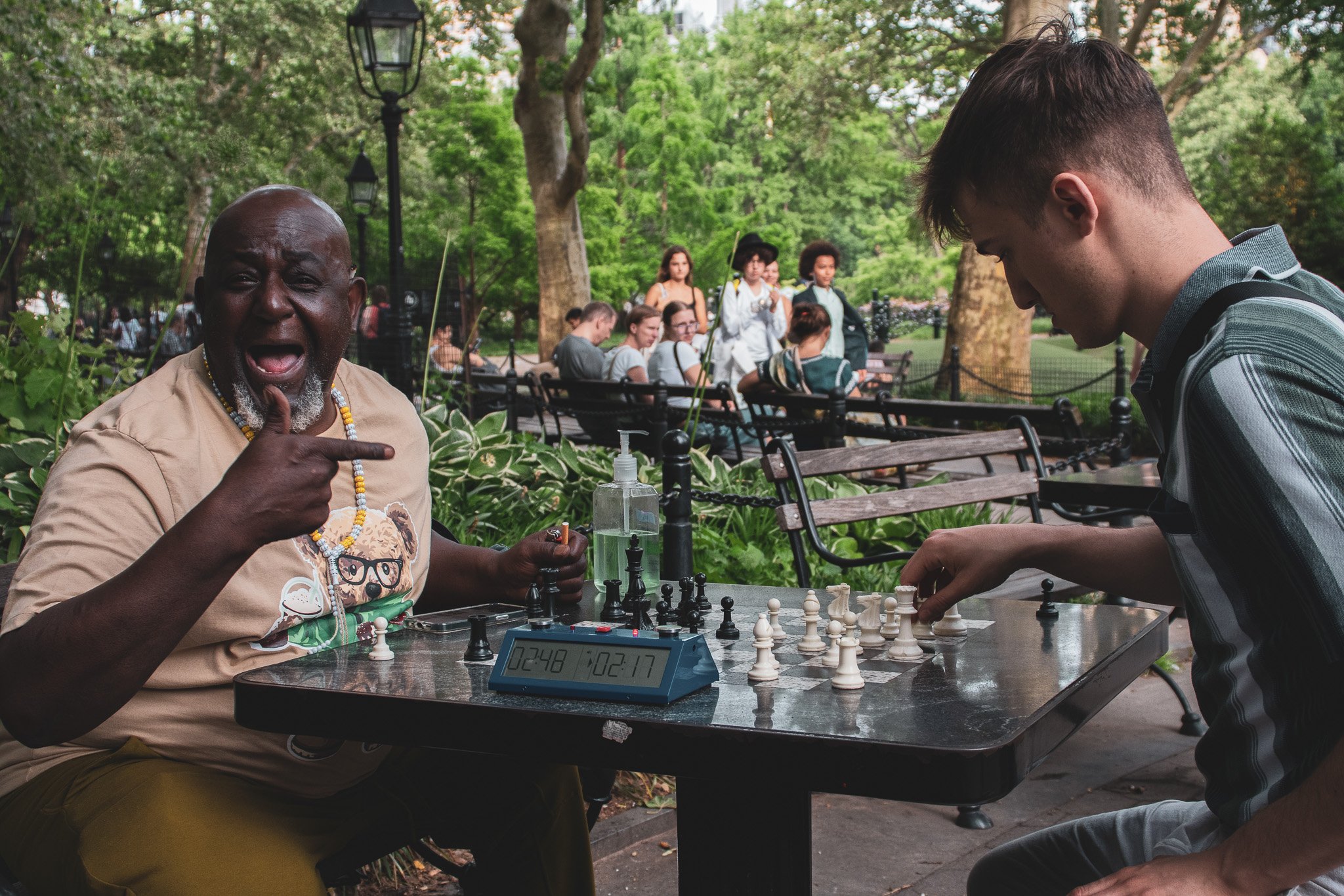 An elderly man with a gray beard and wearing a beige shirt gestures towards a young man with short brown hair who is playing chess in a park. There are several people in the background, some sitting on benches and others standing, surrounded by green