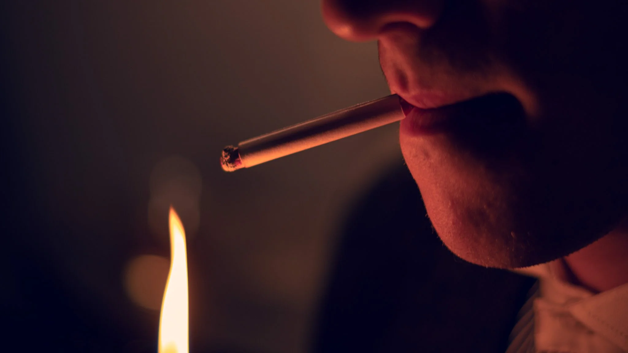 Close-up of a person lighting a cigarette with a match, with a flame visible in the background.