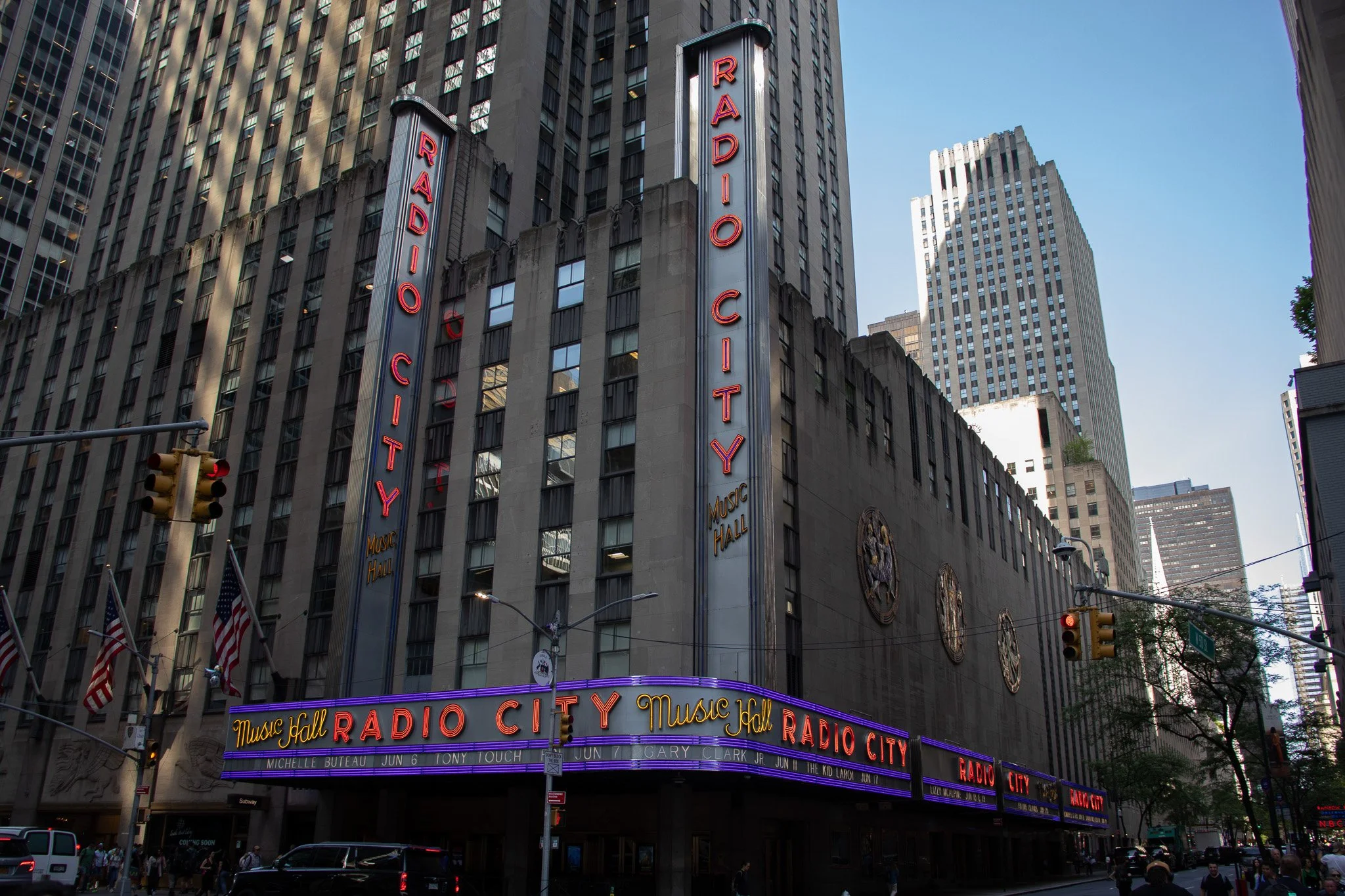 The Radio City music hall building in New York City with tall skyscrapers in the background, illuminated marquee signs, and American flags at street level.