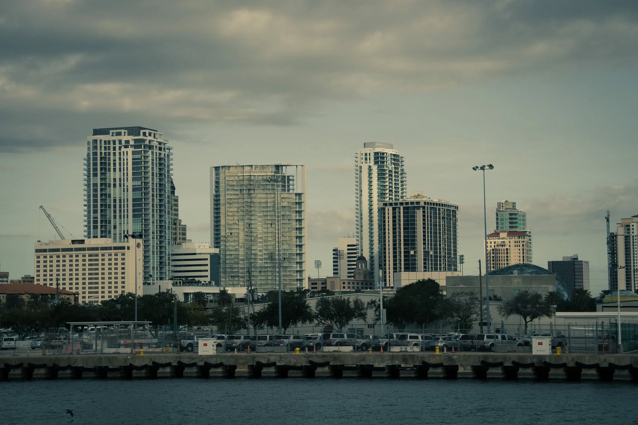 A city skyline with tall modern buildings and a parking lot in the foreground, viewed over a body of water under a cloudy sky.