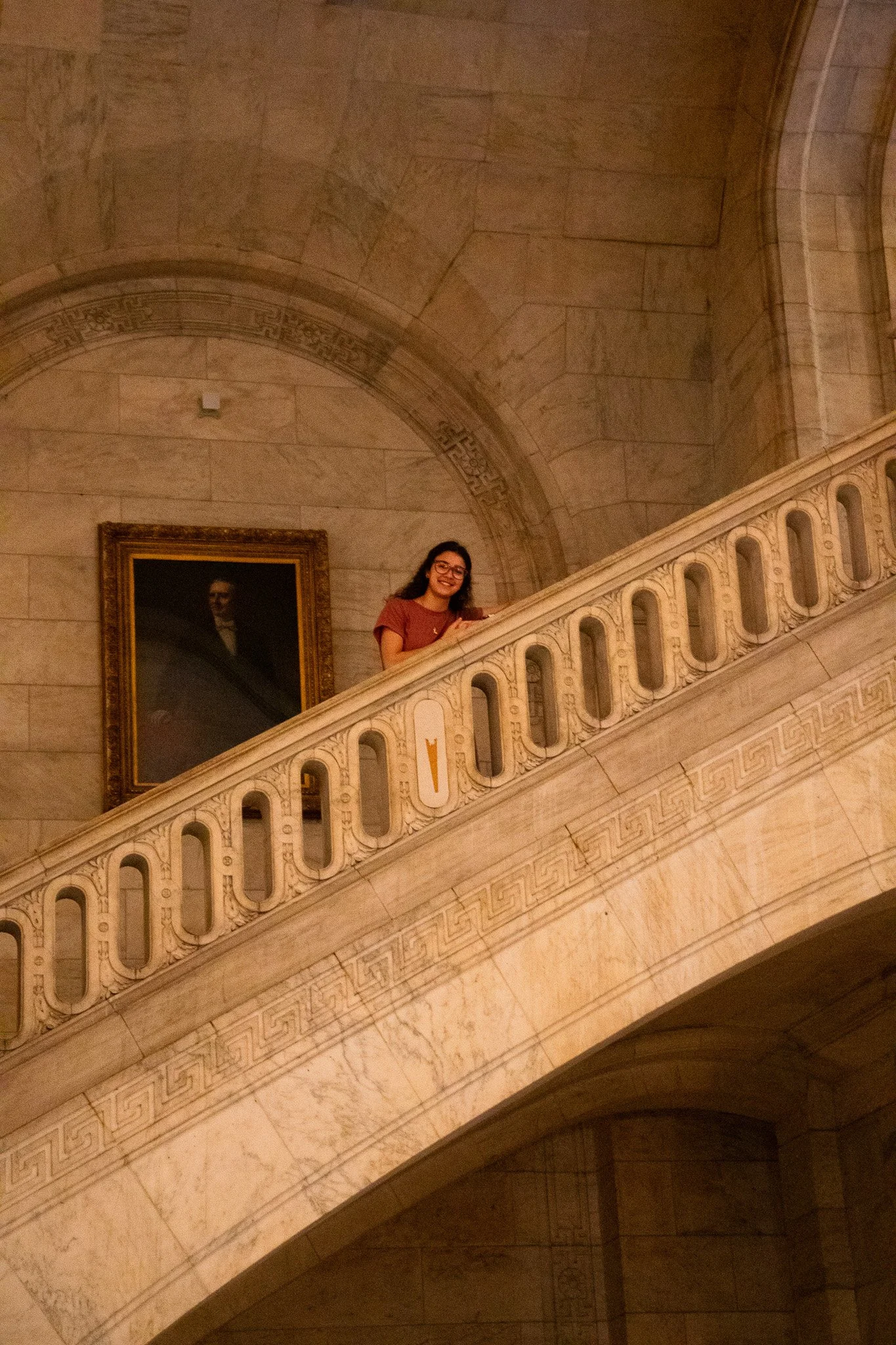 Young woman with glasses and a brown shirt leaning on a decorative marble staircase railing inside a building with beige marble walls and a portrait painting hanging on the wall.