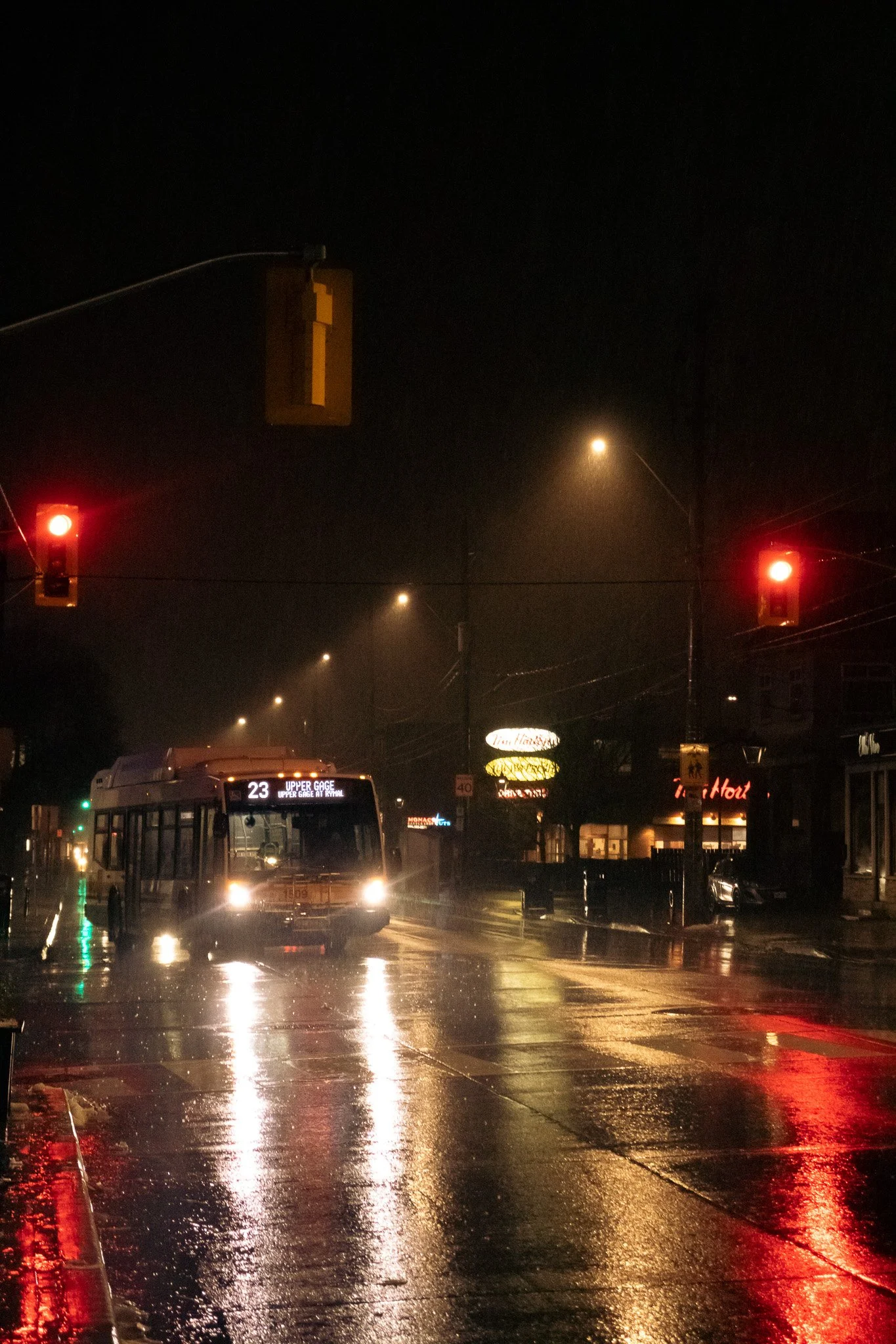 A city street intersection at night, featuring traffic lights with red signals, a bus on the road, and wet pavement reflecting streetlights and signs. The scene looks rainy and foggy, with illuminated signs and street lamps.