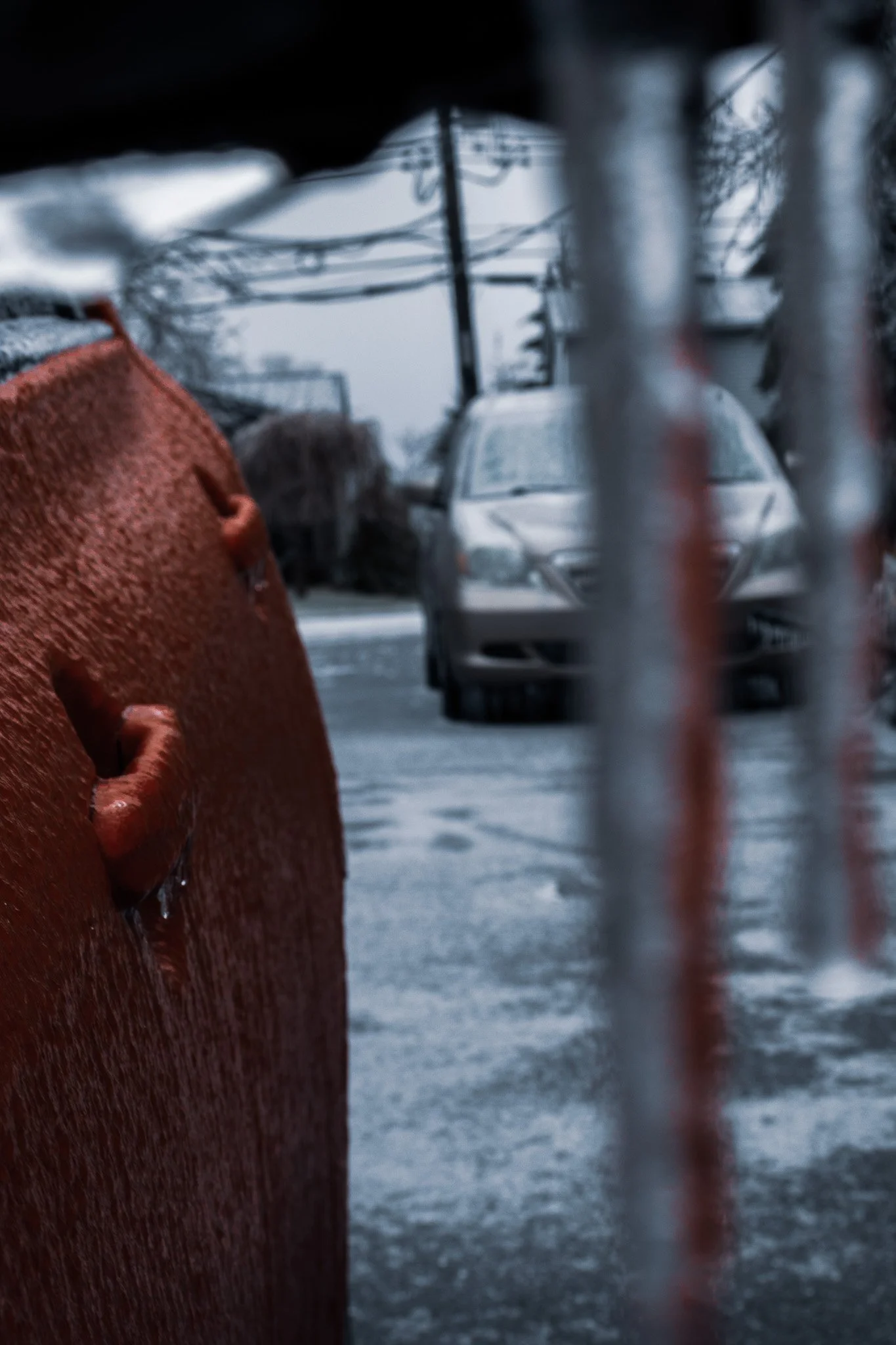 A snow-covered scene seen through a chain-link fence, with a silver car and utility poles in the background, and a person wearing a bright orange glove in the foreground.
