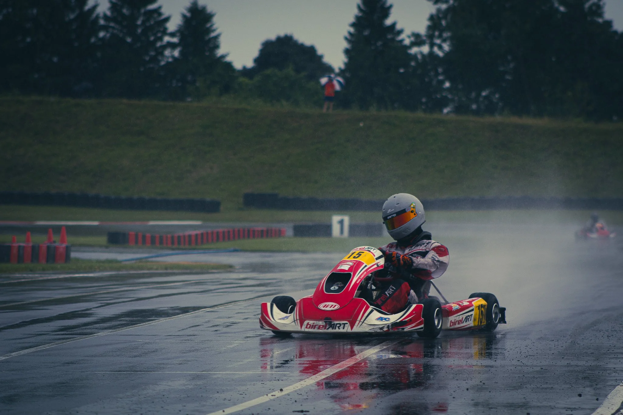 A race car driver in a white helmet and racing suit driving a red and white race car on a wet track during a rainy day. In the background, a spectator with an umbrella stands on a grassy hill with trees.