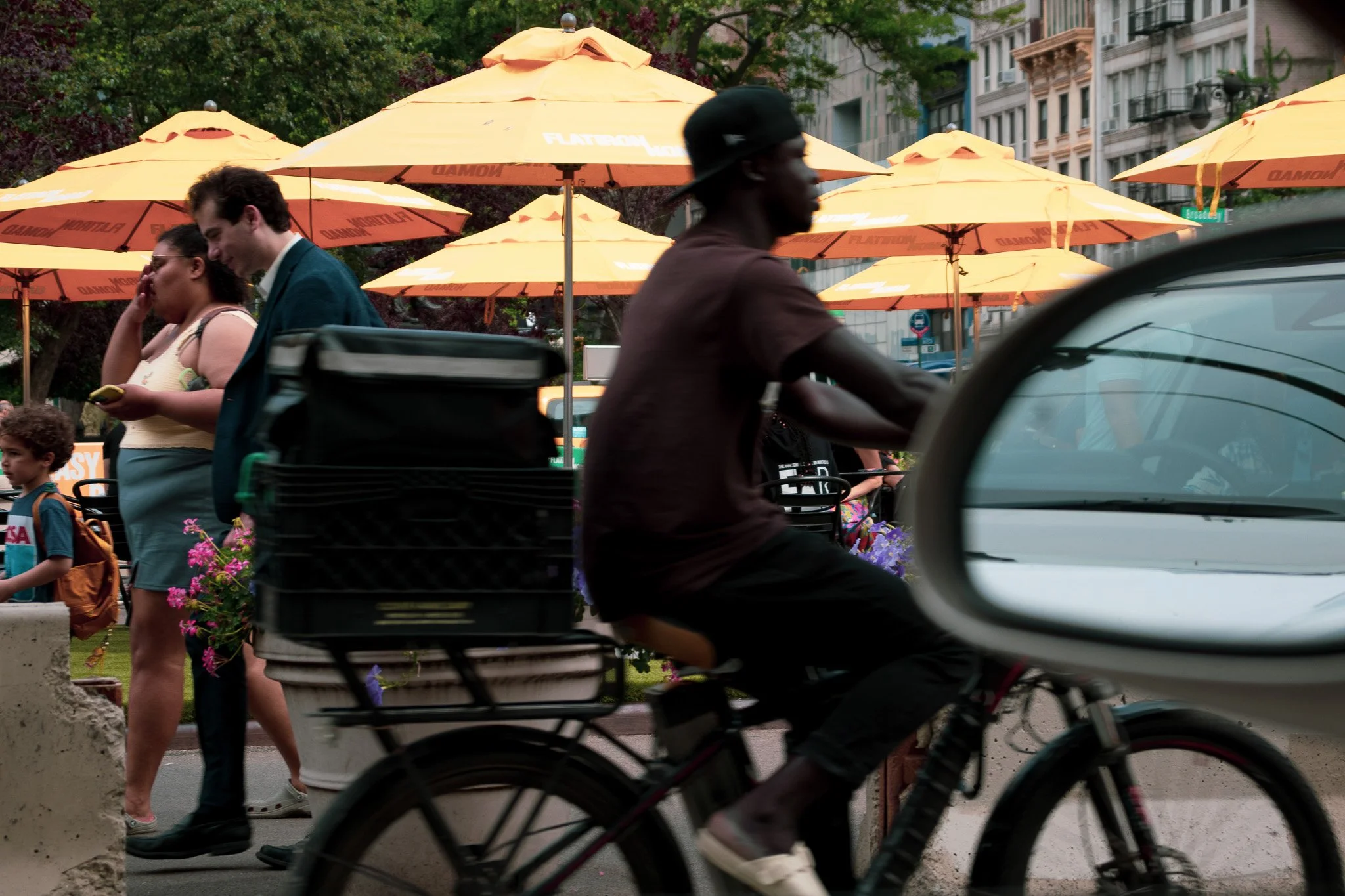 A person riding a bicycle in motion in front of an outdoor seating area with yellow umbrellas and people walking in an urban setting.