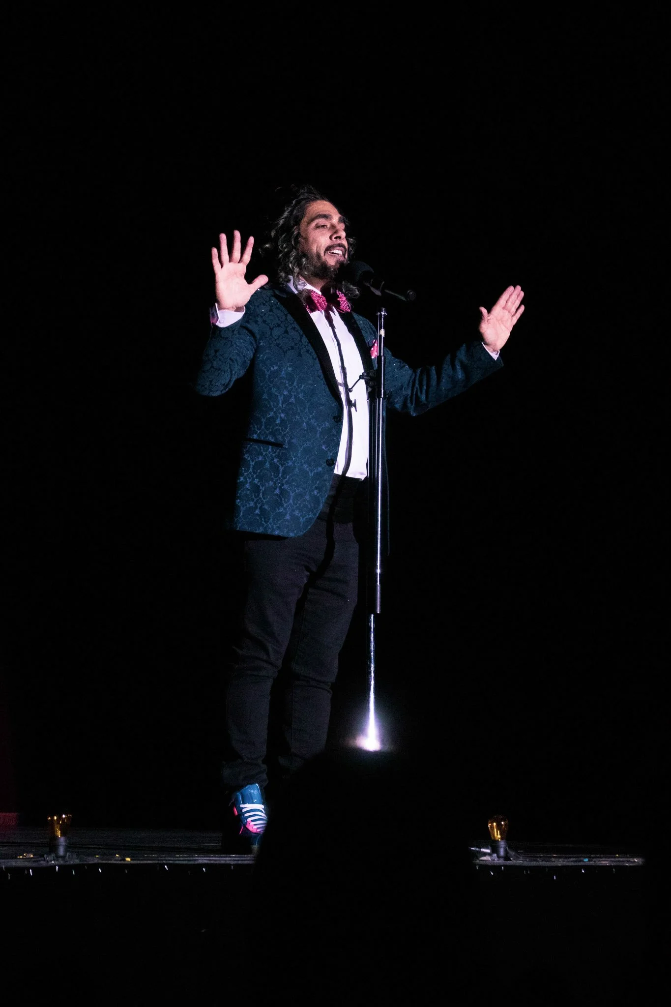 Performer on stage with arms raised, dark background, spotlight on feet, wearing a dark tuxedo with a pink bow tie and colorful sneakers, speaking into a microphone.