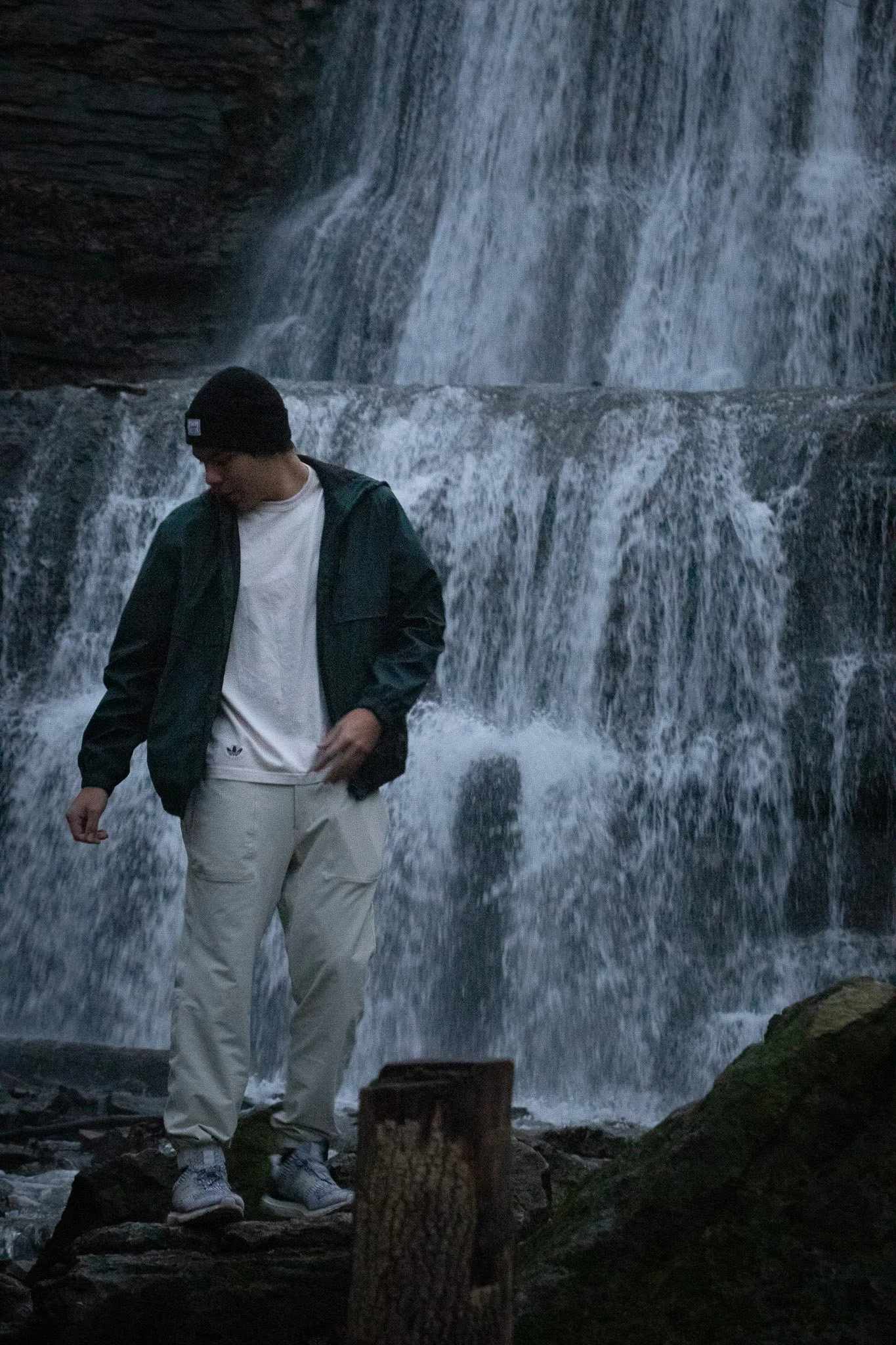 A man wearing a black beanie, green jacket, white t-shirt, and beige pants standing in front of a waterfall at dusk.
