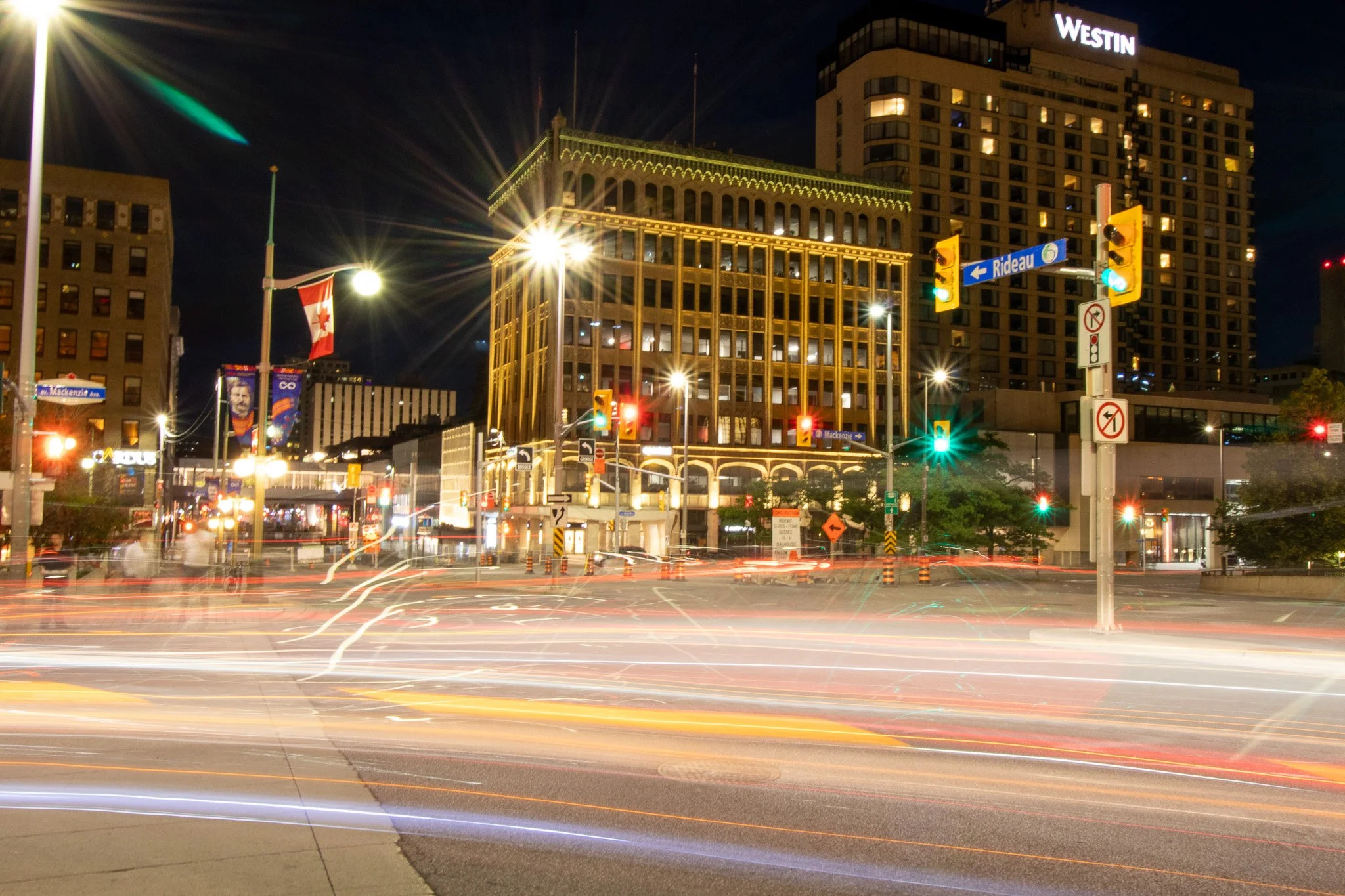 Nighttime city street scene with light trails from moving vehicles, bright streetlights, traffic signals, and tall buildings including a hotel with a "Westin" sign.
