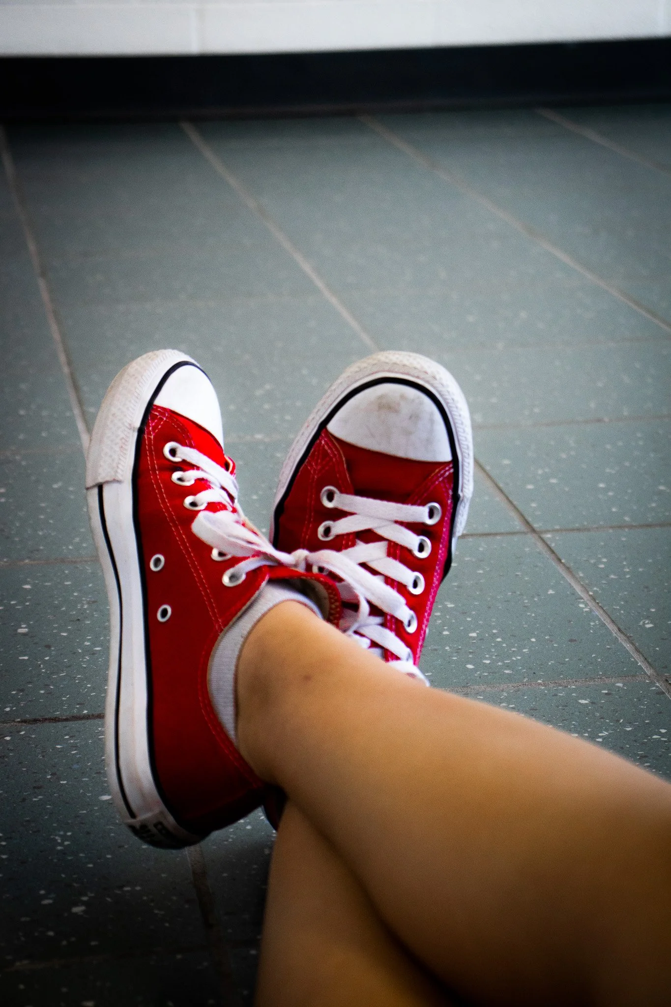 Person sitting on the floor wearing red sneakers with white laces, with their legs extended and crossed at the ankles, on a dark tiled floor.