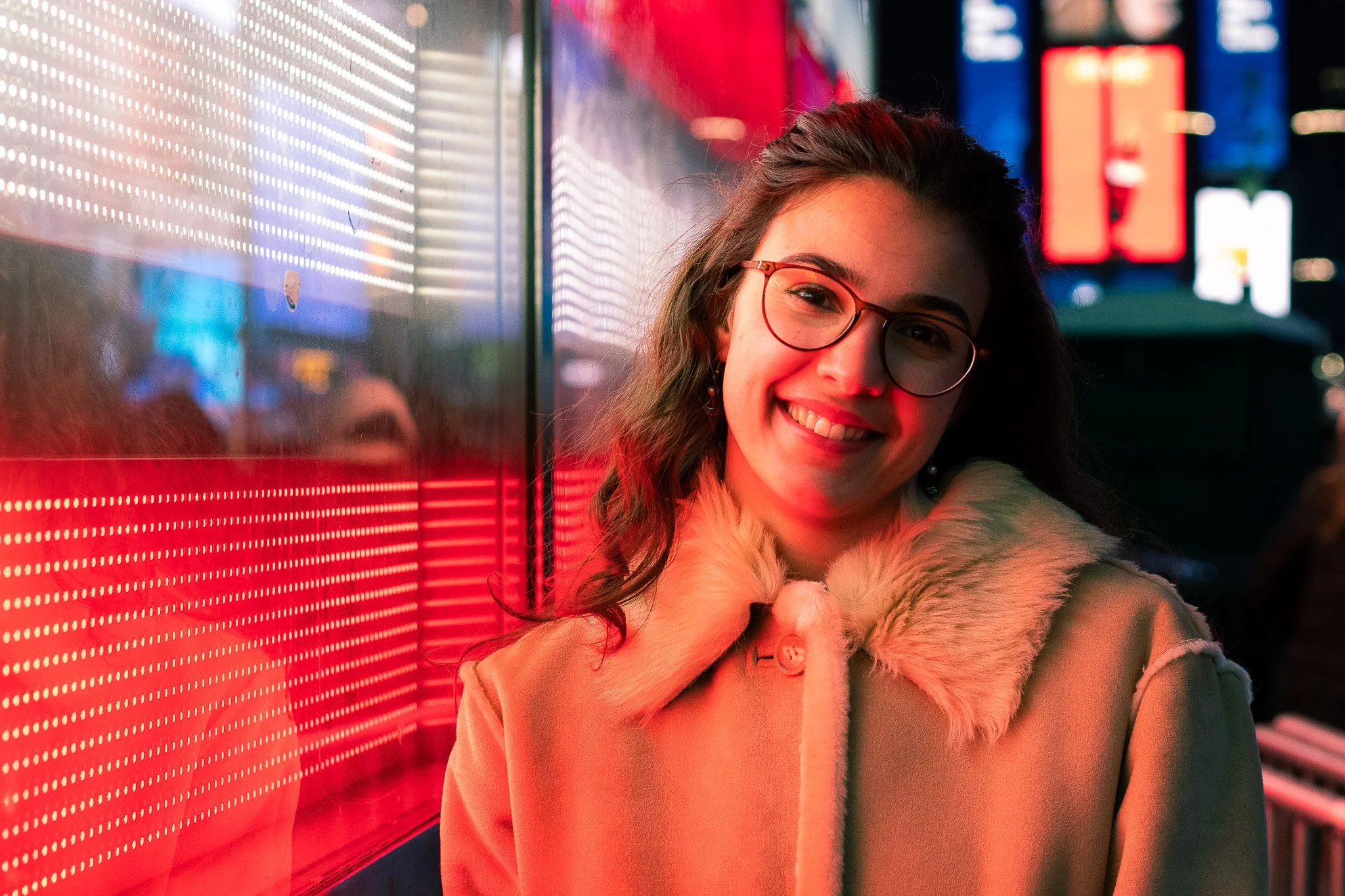 A smiling young woman with glasses and earrings, wearing a beige coat with a fur collar, standing next to a brightly lit, colorful, illuminated digital display at night.