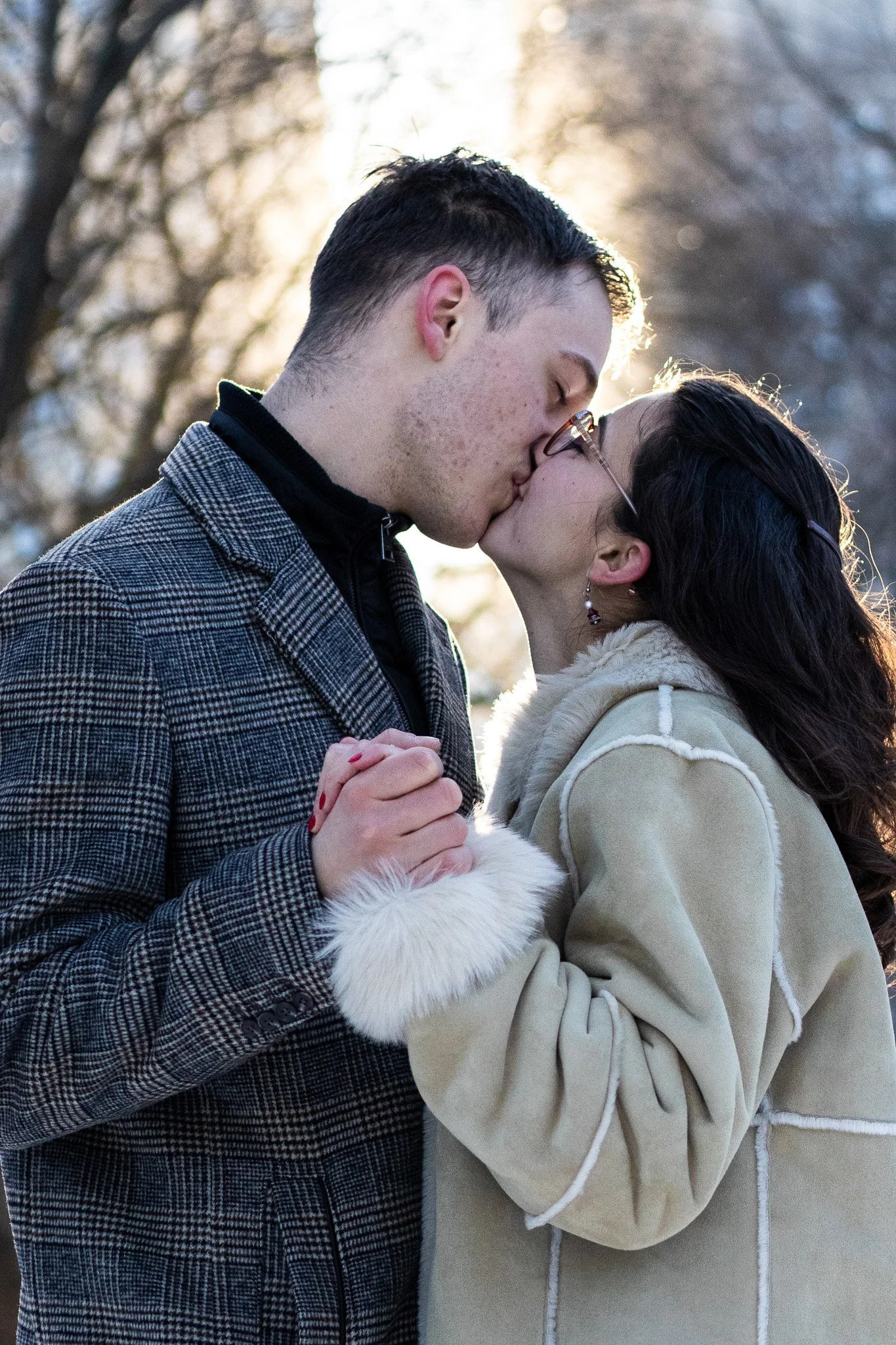 A couple shares a kiss outdoors during winter, holding hands, with bare trees and sunlight in the background.