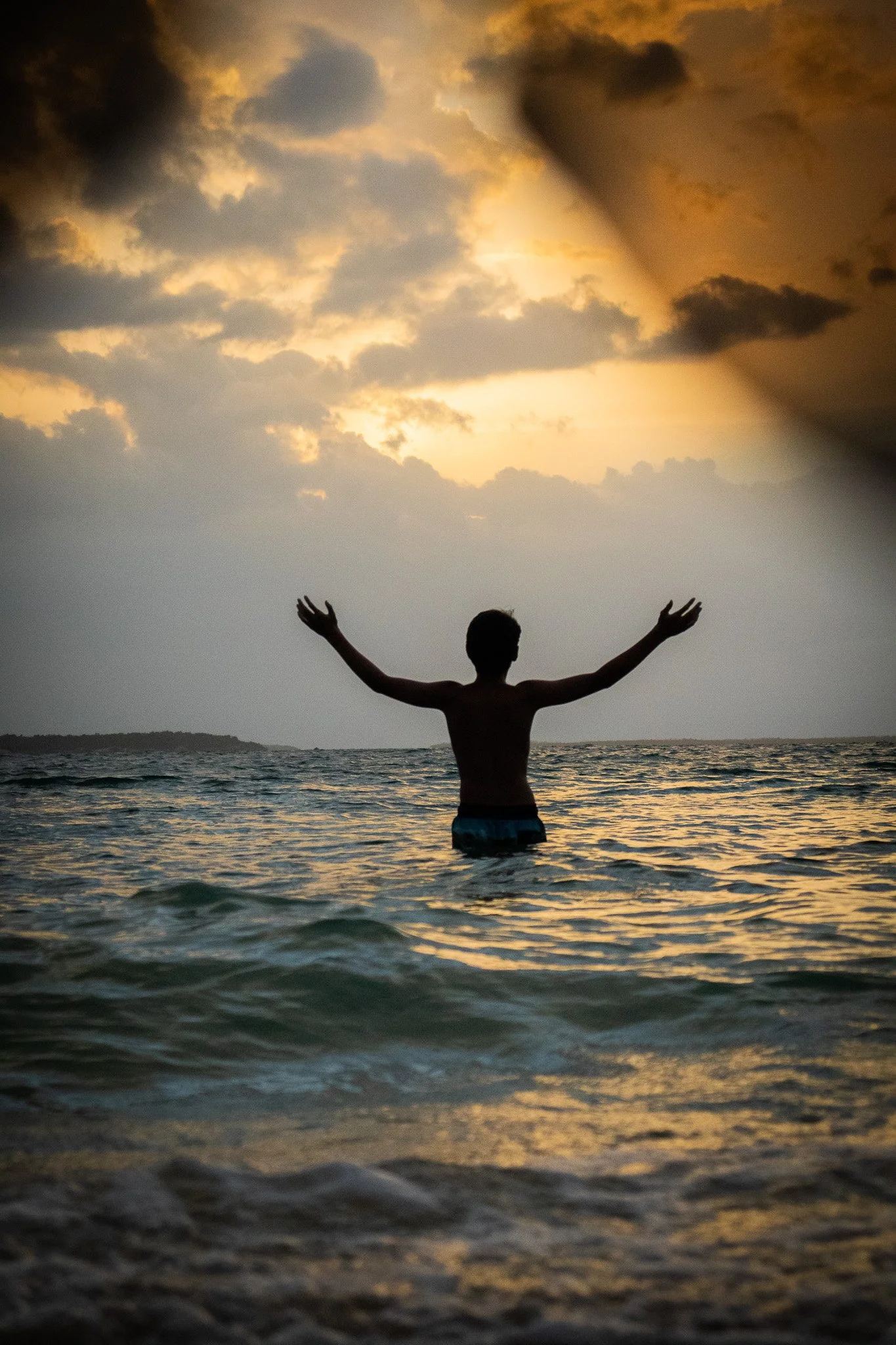 Person standing in the ocean with arms raised at sunset or sunrise, dark clouds in the sky, water waves, and a distant shoreline.