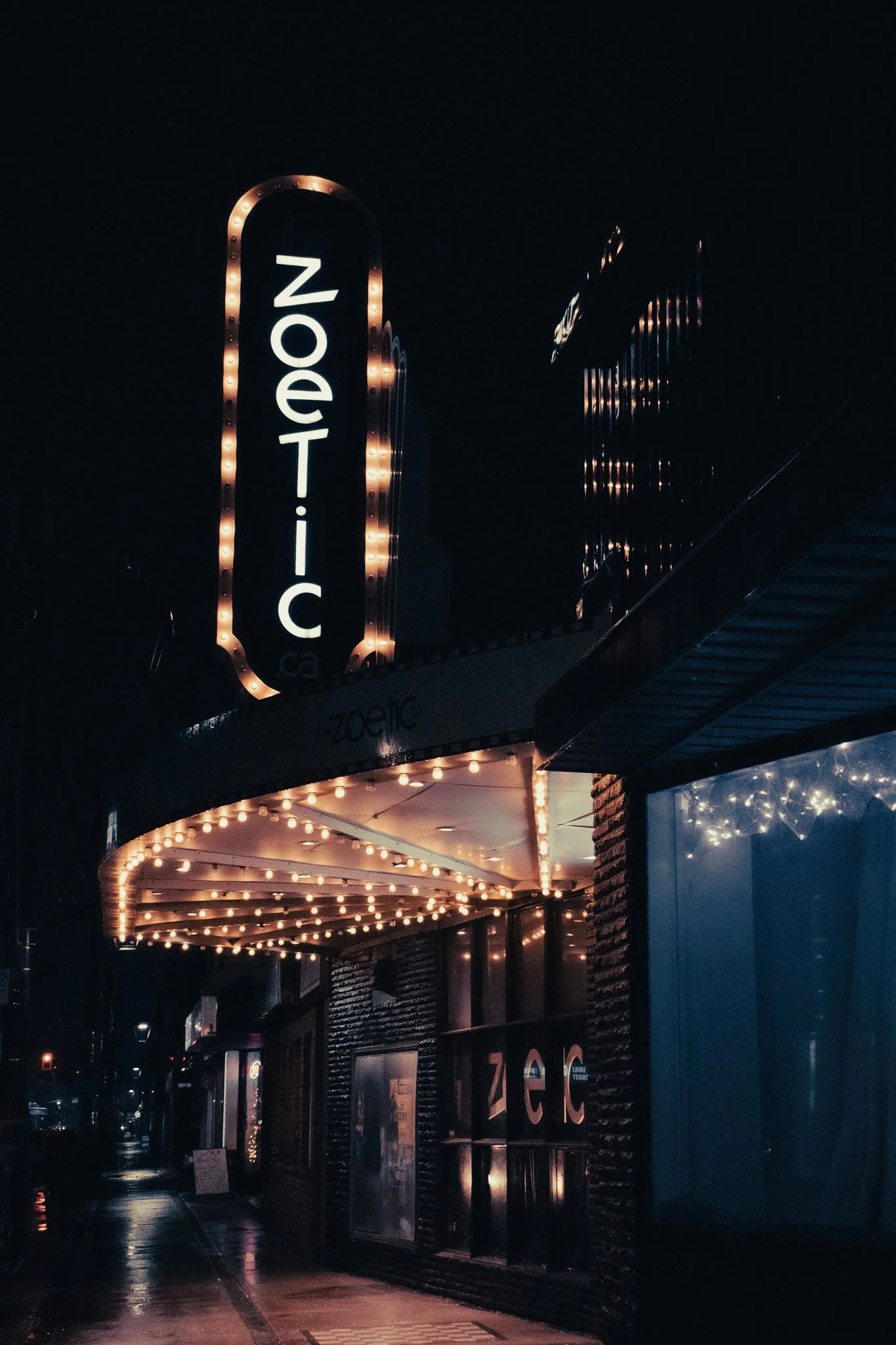 Night view of a theater or music venue with illuminated marquee and signage. The marquee is decorated with small light bulbs around its edges. The venue name is partially visible as 'ZEC'. The scene appears to be on a city sidewalk.