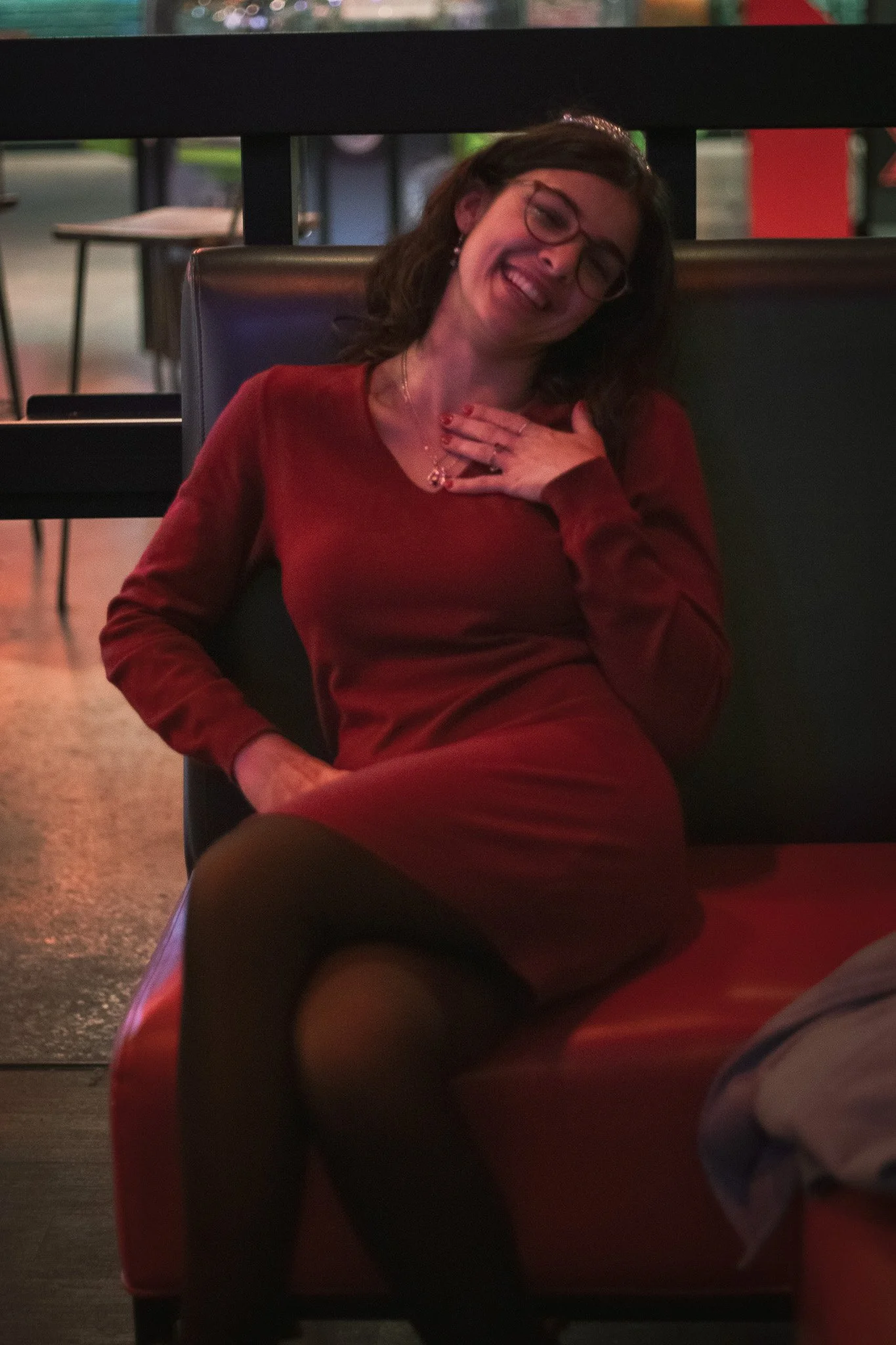 A woman wearing glasses and a red dress, with dark wavy hair, smiling and touching her chest with her hand, sitting on a black chair in a dimly lit restaurant or cafe.
