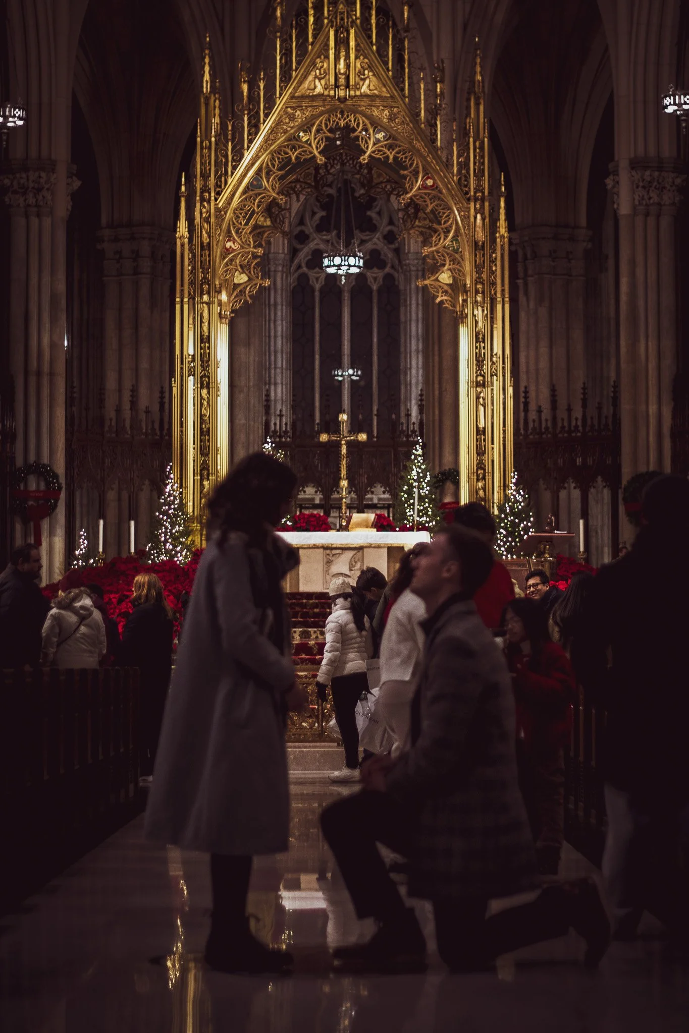 Inside a church decorated for Christmas, with people standing and sitting, and a gold altar with a cross, Christmas trees, and poinsettias.