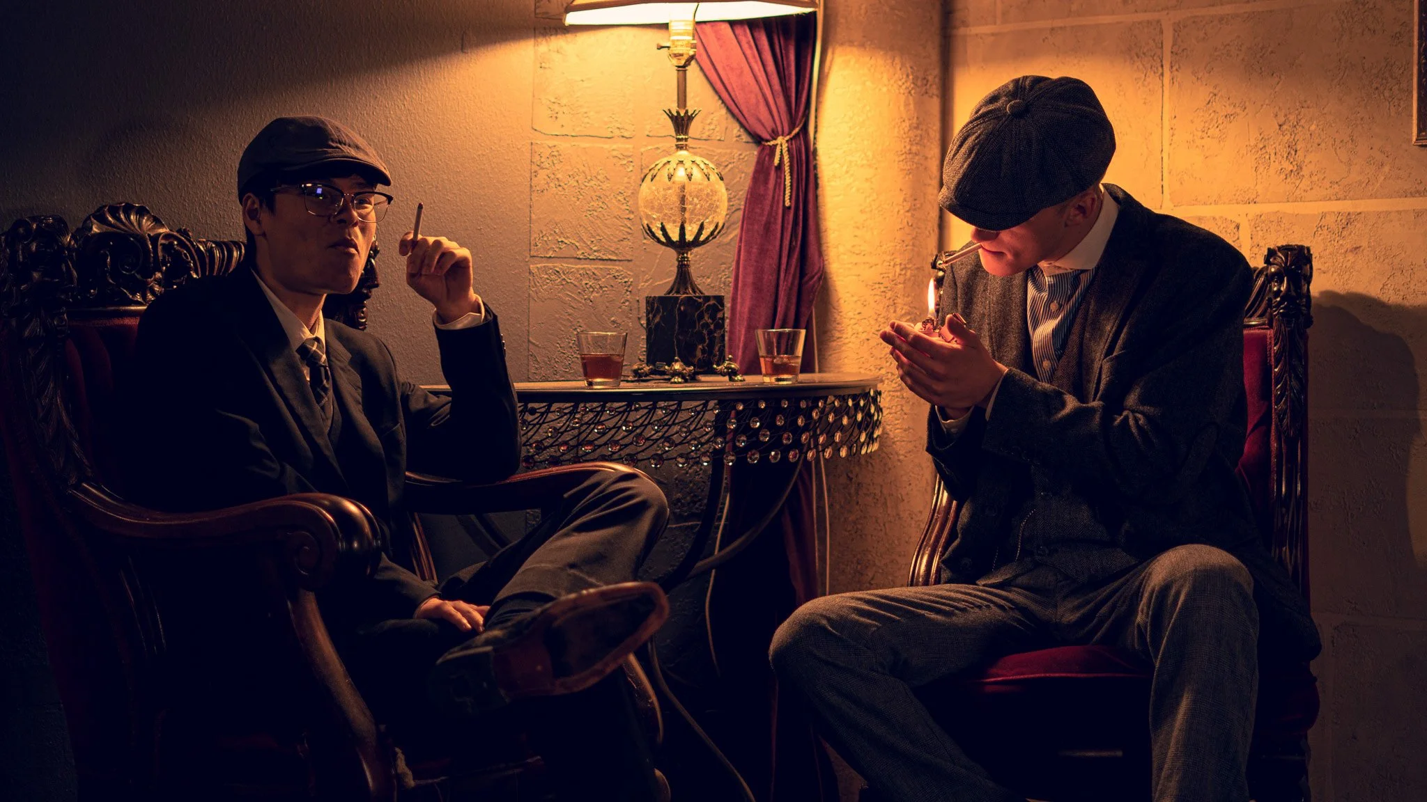 Two men in vintage-inspired suits and hats sitting in a dimly lit room. One is smoking a cigarette and the other is looking at a phone. There is a table with drinks and a decorative lamp between them.