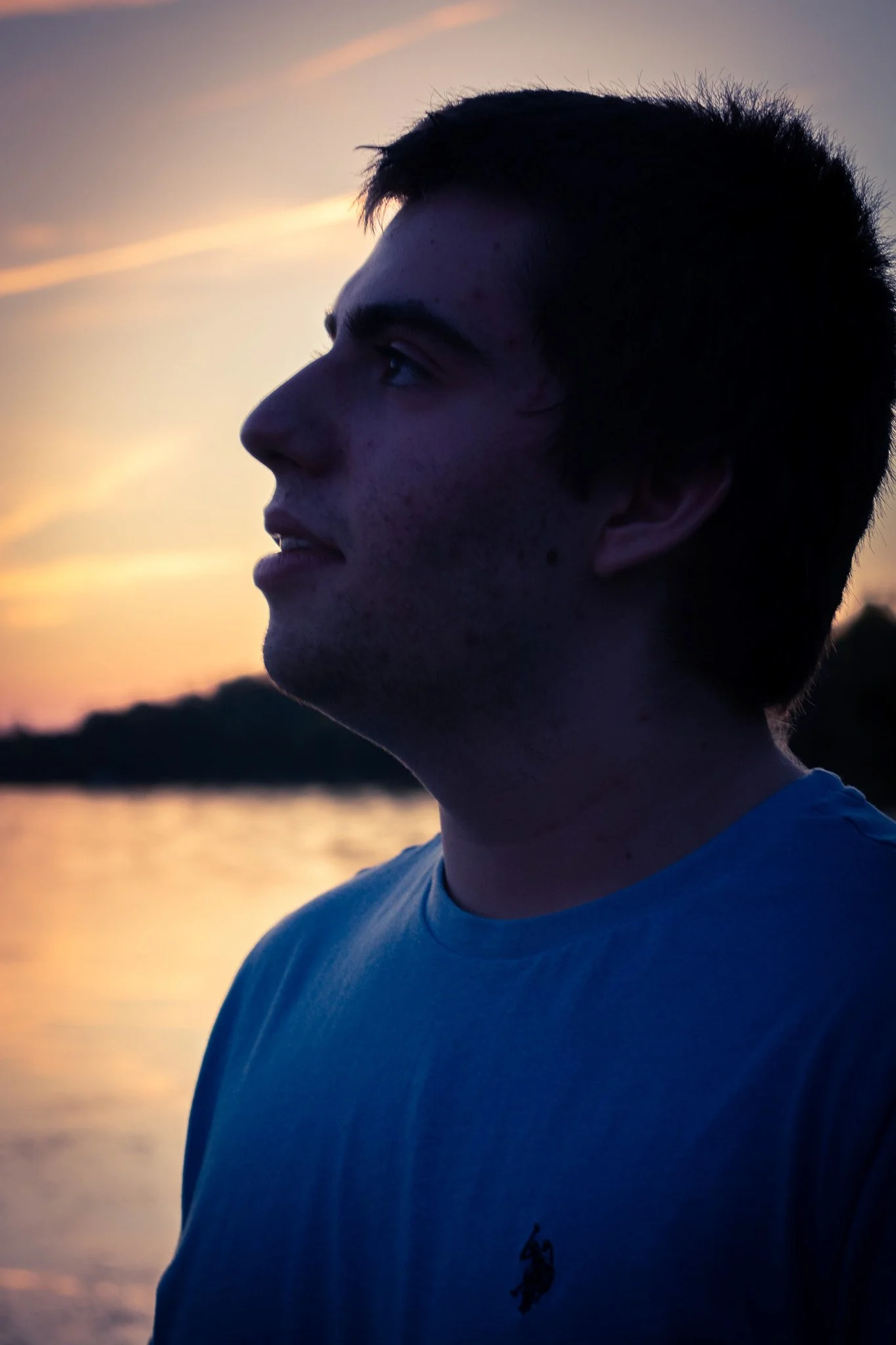 Profile of a young man with short dark hair wearing a blue t-shirt with a small logo, looking to the left against a sunset over a body of water.