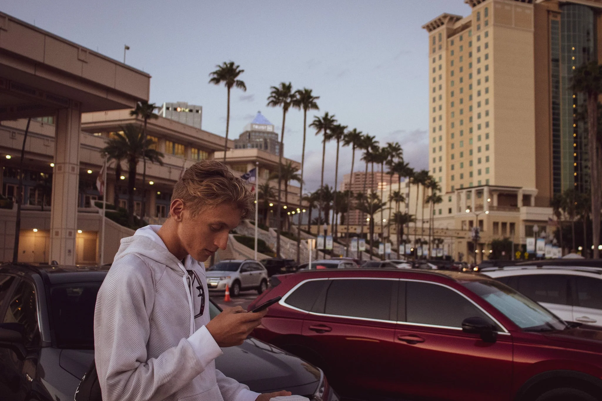 A young man with light brown hair looking at his phone, wearing a light-colored hoodie, standing next to a red car in a parking lot with tall palm trees and a city skyline in the background during dusk.