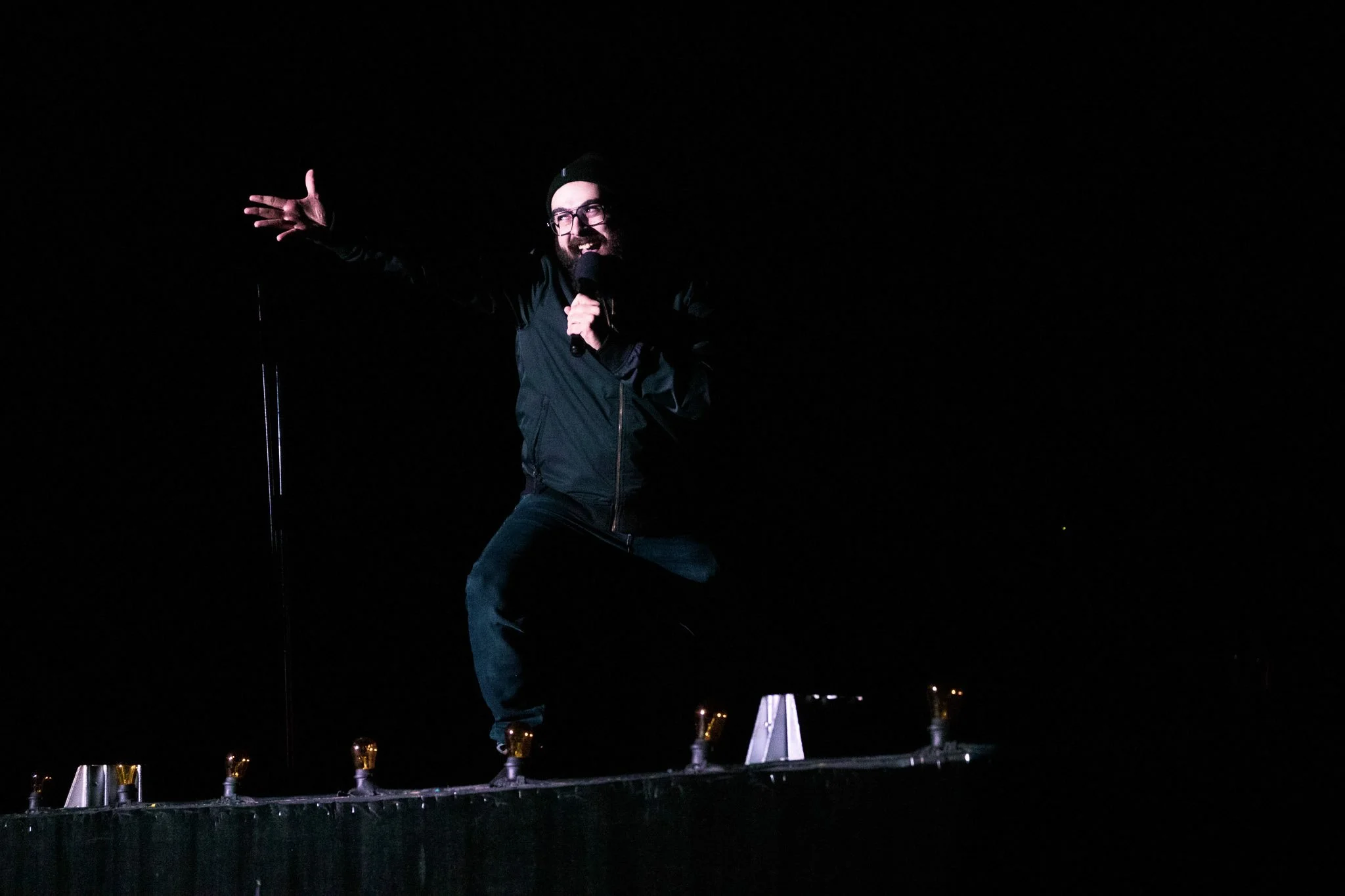 A man with glasses and a beard, dressed in black, performing stand-up comedy at night with a microphone in hand, standing on a dark stage.