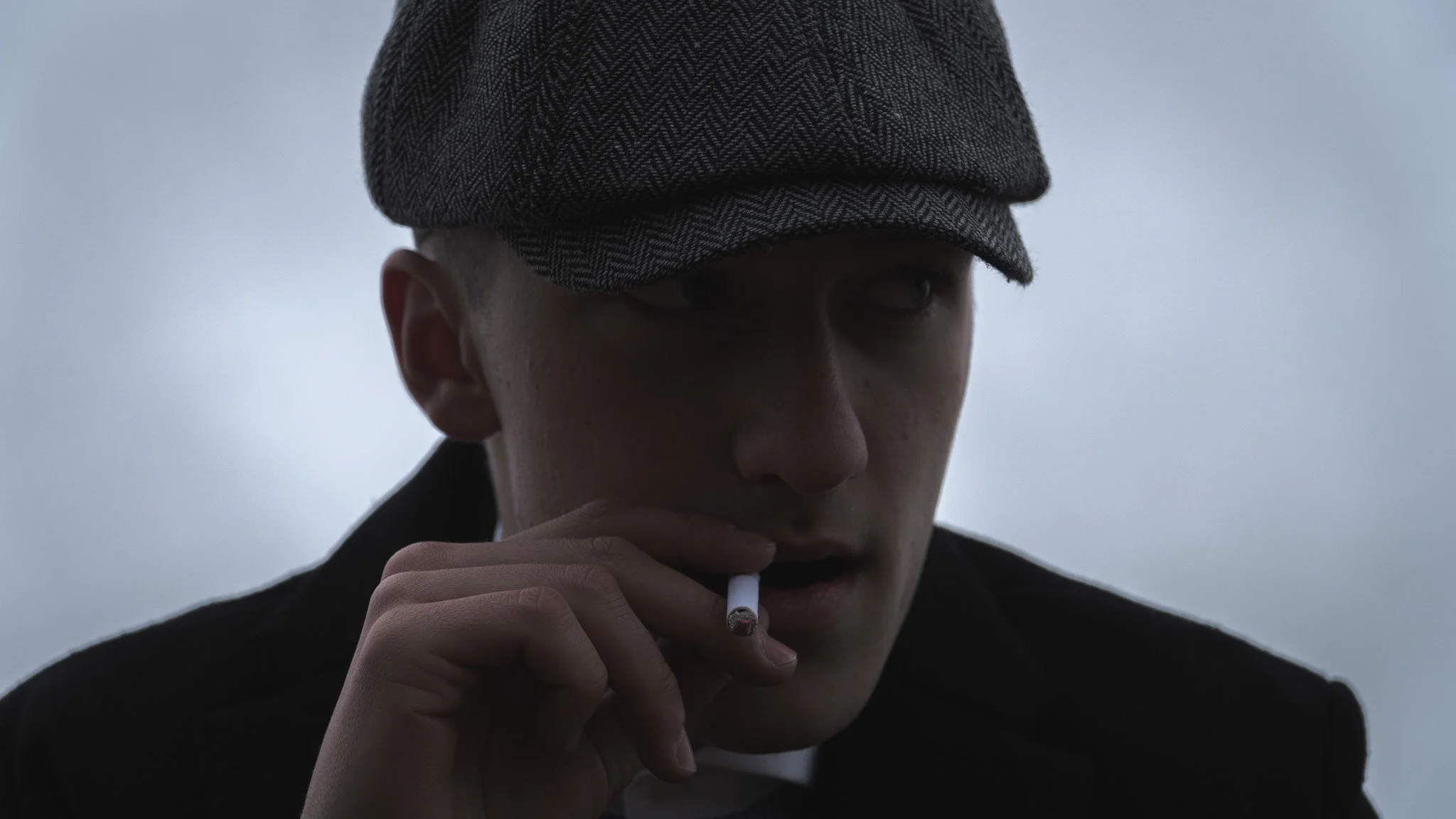 A person wearing a grey herringbone cap and black jacket, holding a cigarette in hand, with a serious expression and touching their lips with fingers.