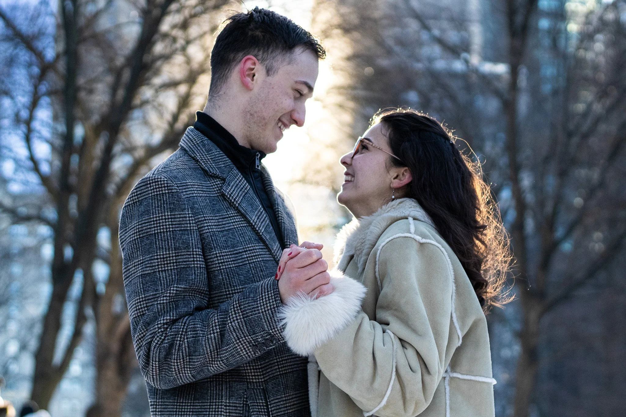A young man and woman are standing close together outdoors, smiling and holding hands, with trees and sunlight in the background.