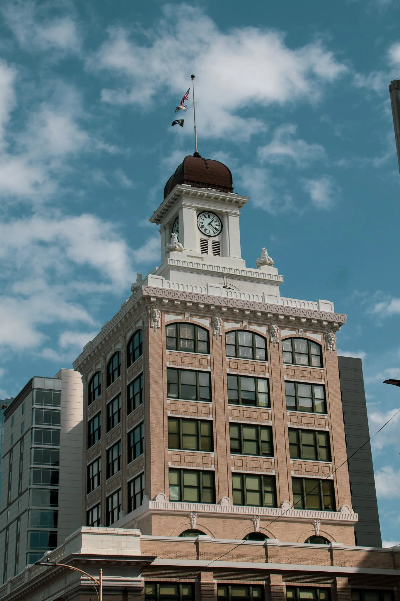 A tall building with a clock tower, featuring a brown roof and white decorative elements, with flags flying on top against a partly cloudy sky.