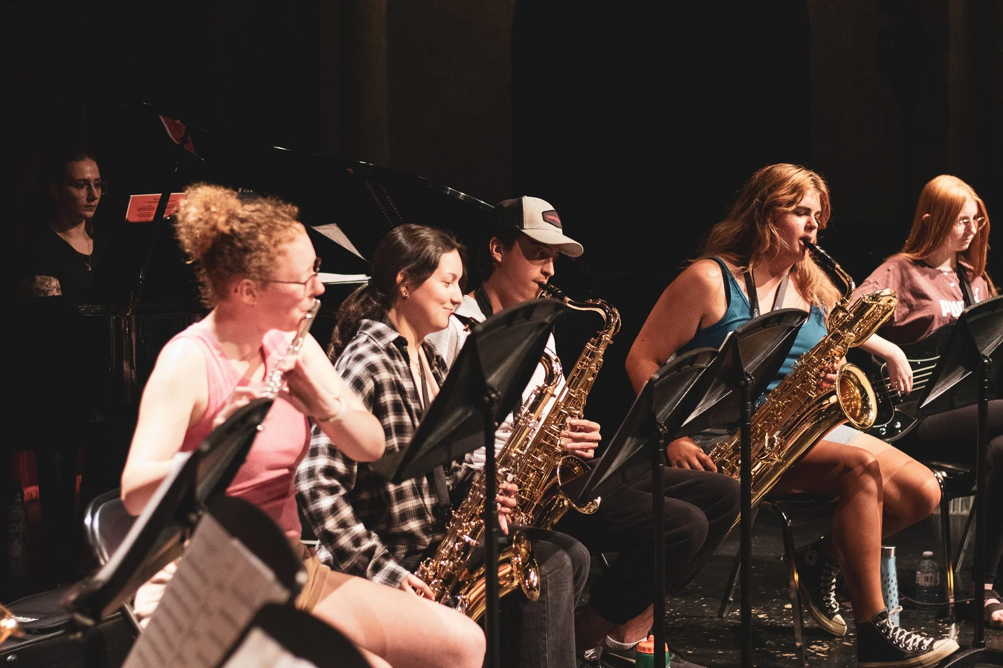 A group of young women and men playing saxophones on a stage during a concert, with a black grand piano in the background.