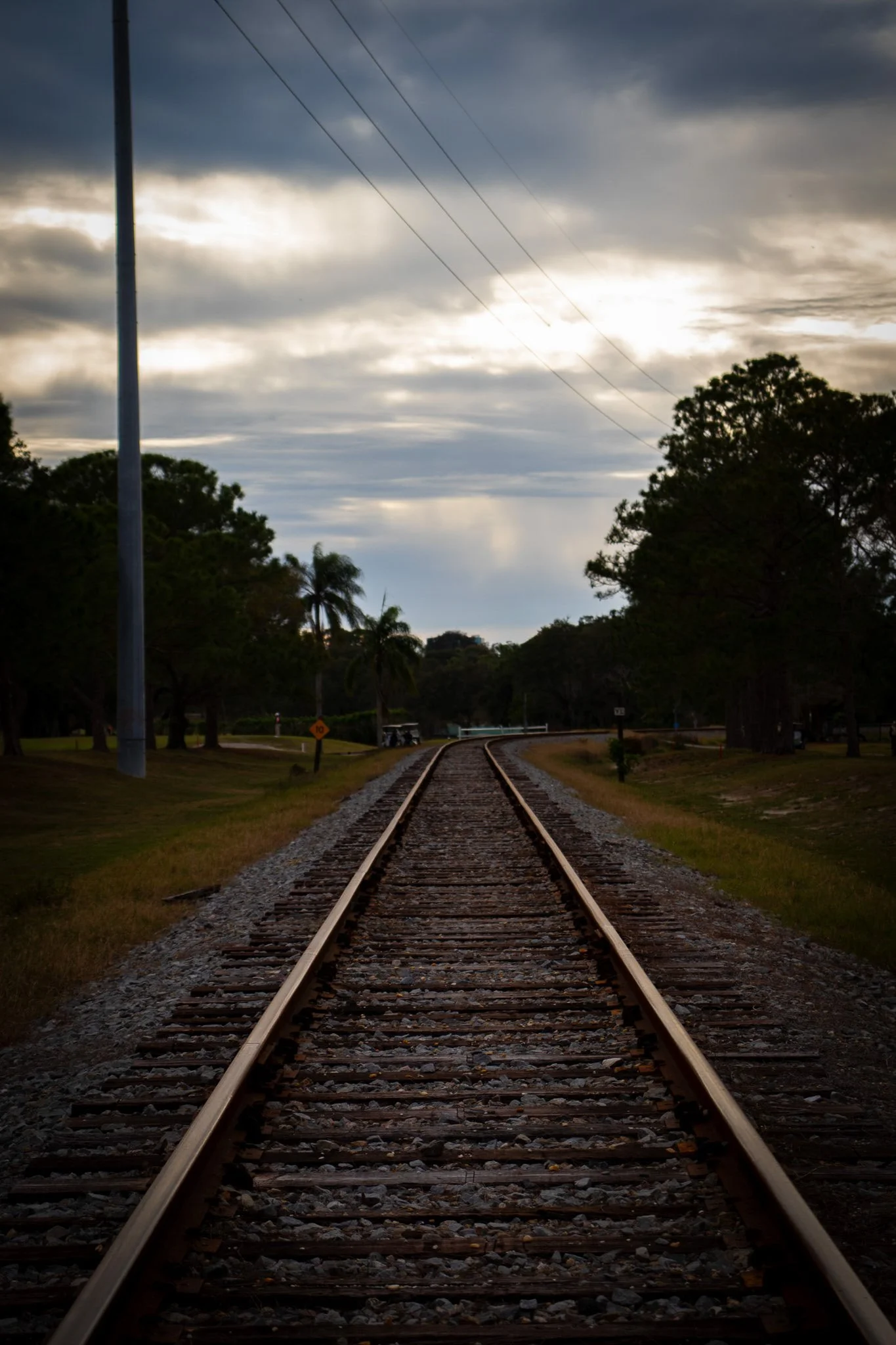 A straight railway track extending into the distance, surrounded by trees and greenery, under a cloudy sky with some sunlight breaking through.