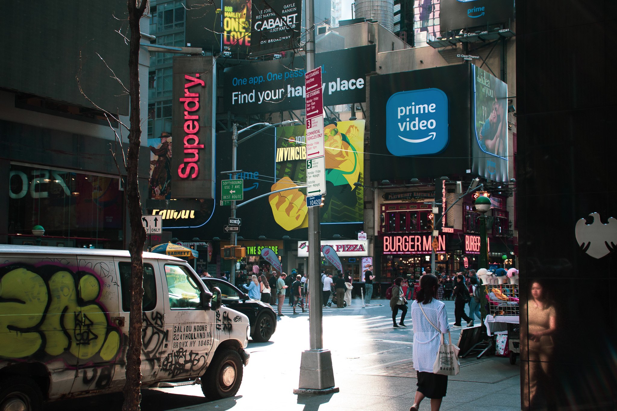 Busy city street scene with pedestrians crossing and standing, billboards and signs, including an Amazon Prime Video advertisement, storefronts like Burger King, and graffitied van parked on the street. Tall buildings surround the area.