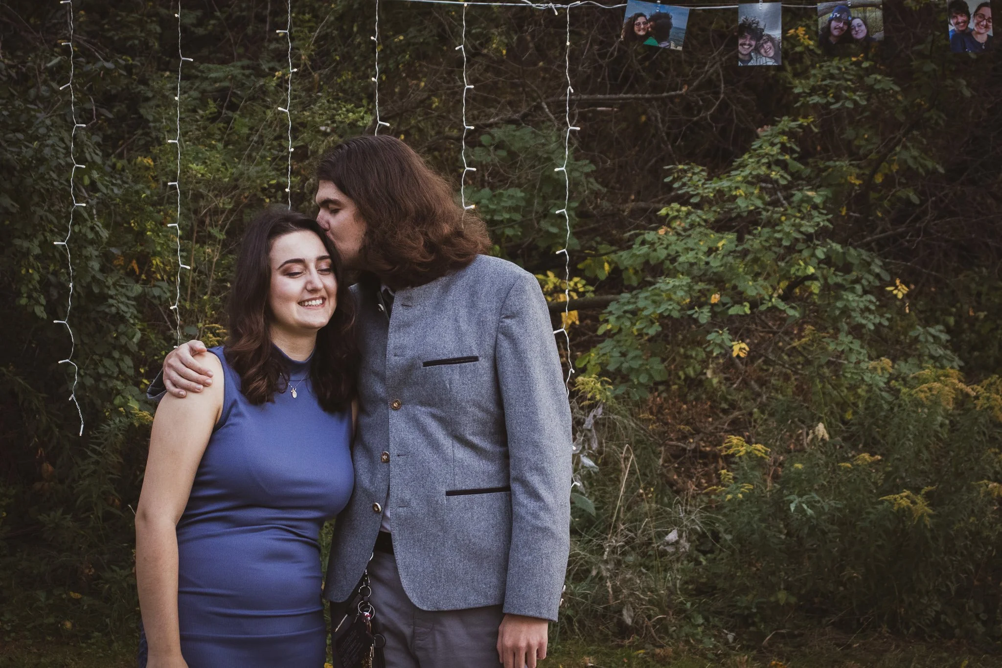 A young woman and a man stand outdoors in front of a backdrop of greenery and string lights, sharing a moment of affection. The woman has long dark hair and is smiling with her eyes closed, while the man has long brown hair and is kissing her on the 