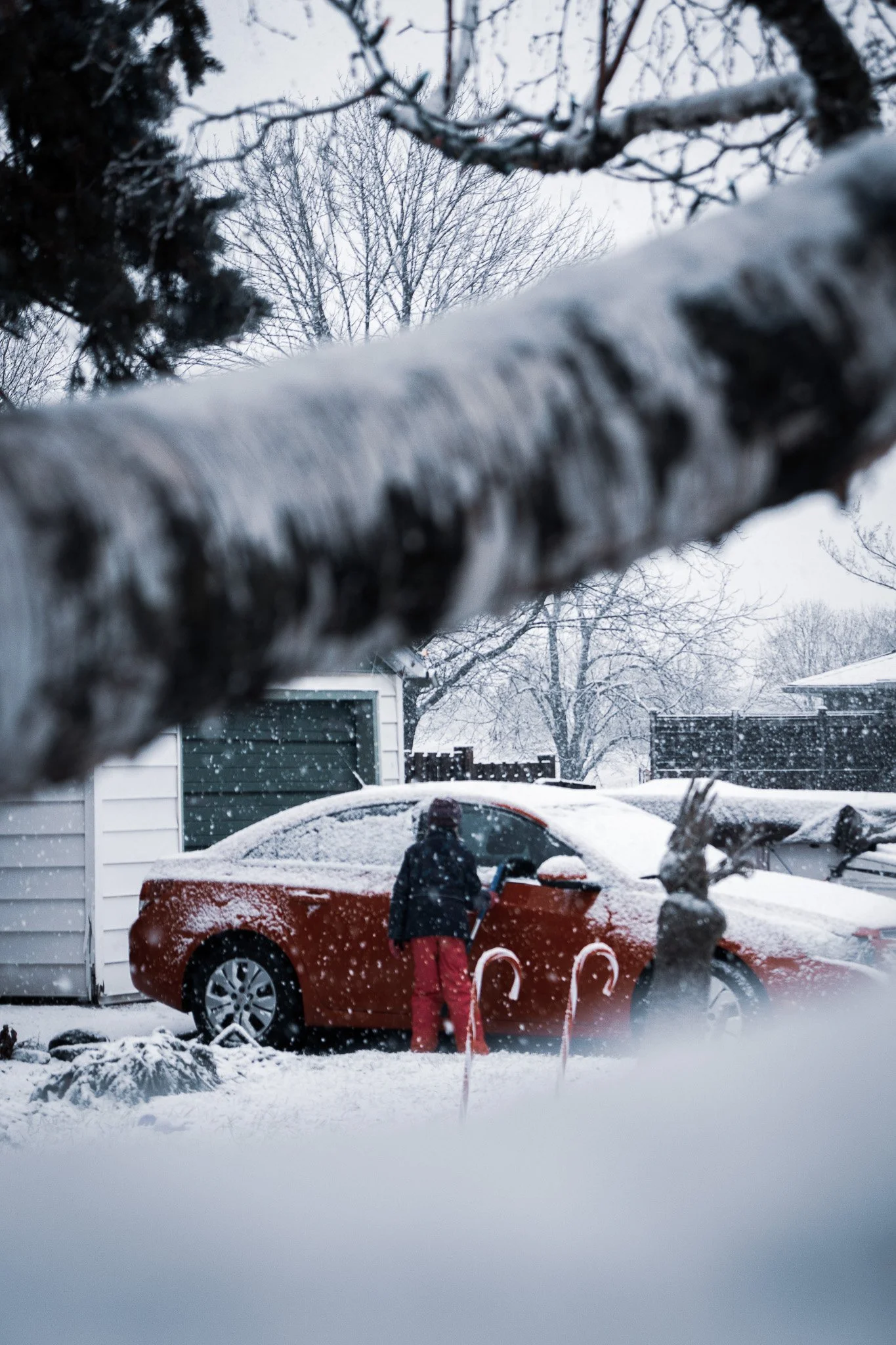 A person in red pants and a dark jacket shoveling snow in a driveway next to a red car, with snow-covered trees and a house in the background during a snowstorm.