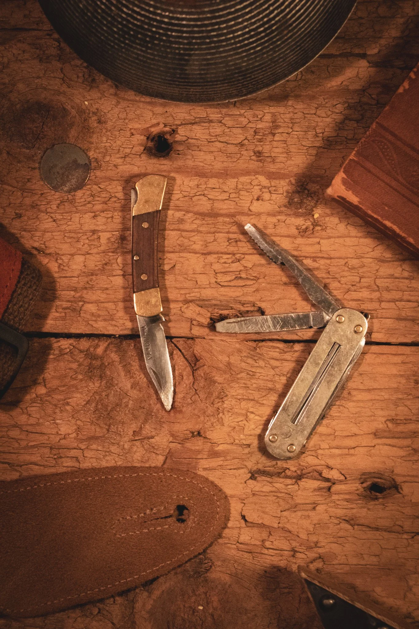 Wooden surface with a folding pocket knife, a serrated folding knife, and a small folding saw, along with a leather wallet and a leather wallet with a button, and a small round metal object.