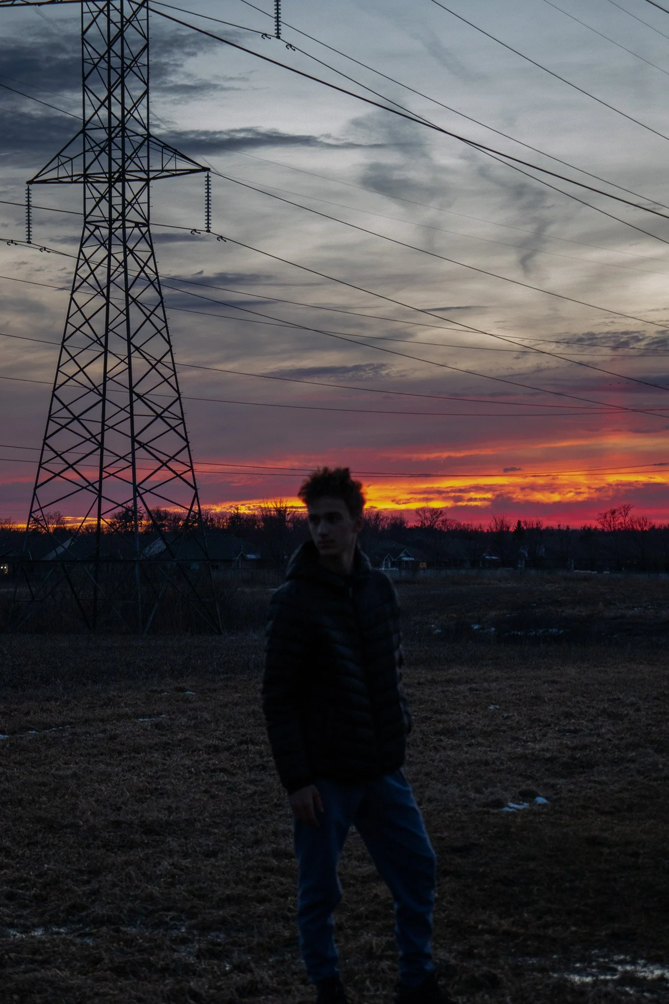 A young man standing outdoors at sunset with power lines and a transmission tower in the background.