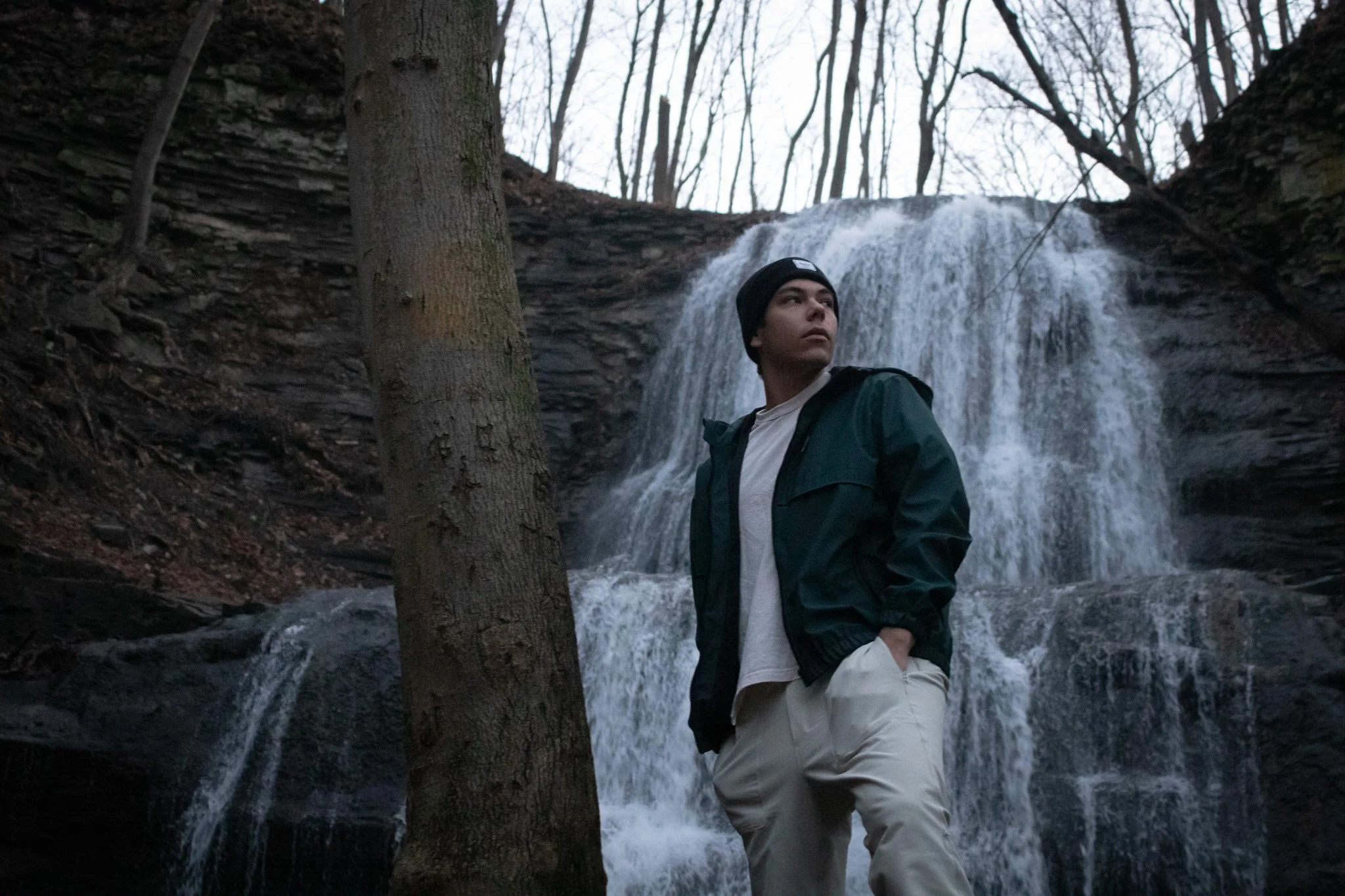 A young man standing in front of a waterfall in a forest during winter, wearing a black beanie, a dark jacket, and light-colored pants.