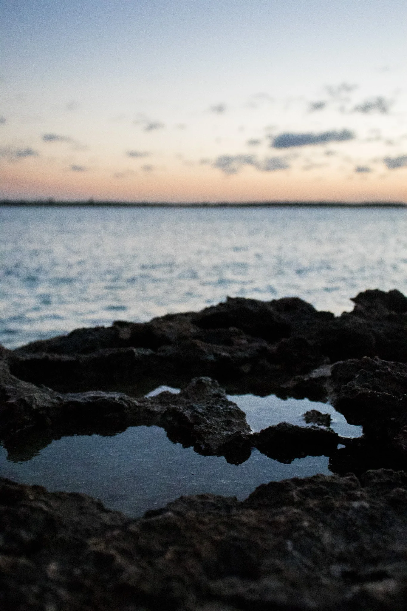 A blurred landscape of a body of water with rocks in the foreground and a sunset sky with clouds in the background.