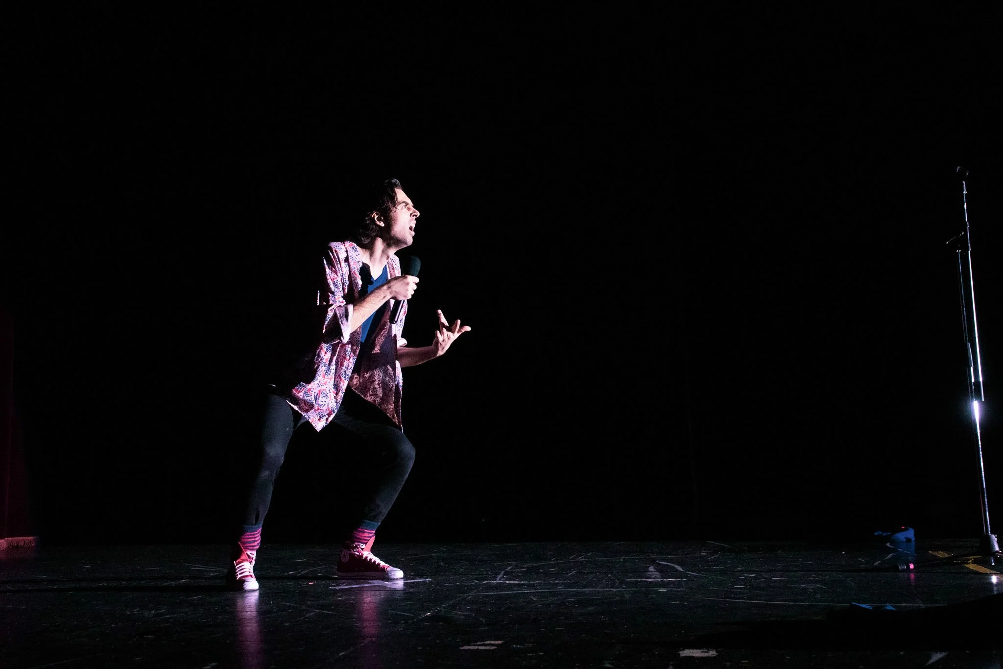A person on stage performing with a microphone, wearing colorful clothing and socks, with a stand and microphone to the right against a black background.
