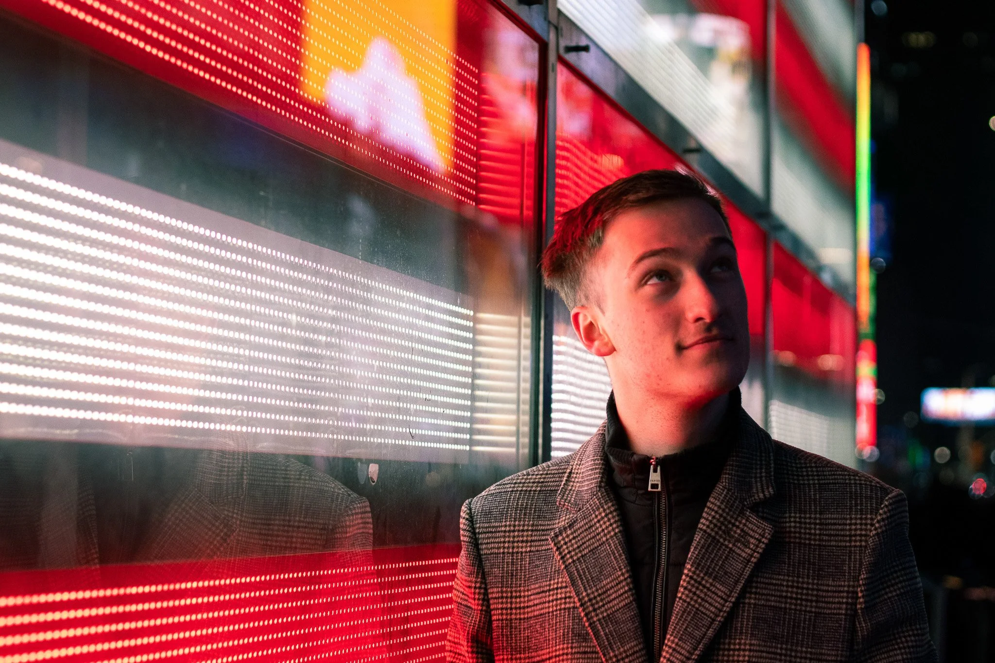 A young man wearing a plaid jacket stands near a brightly lit red and white LED display on a city street at night.