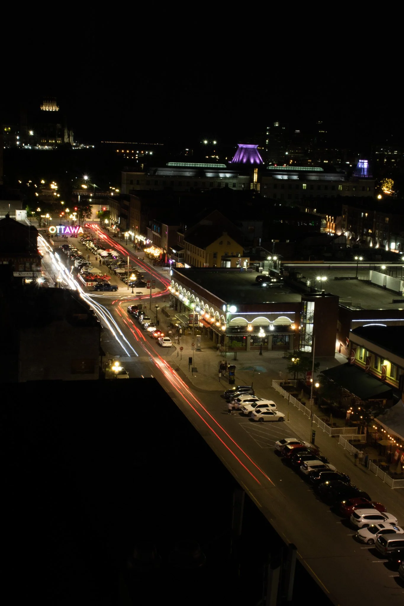 Nighttime cityscape with illuminated buildings and streets, including a bright sign that reads 'OTTAWA' with streaks of car lights on the roads.